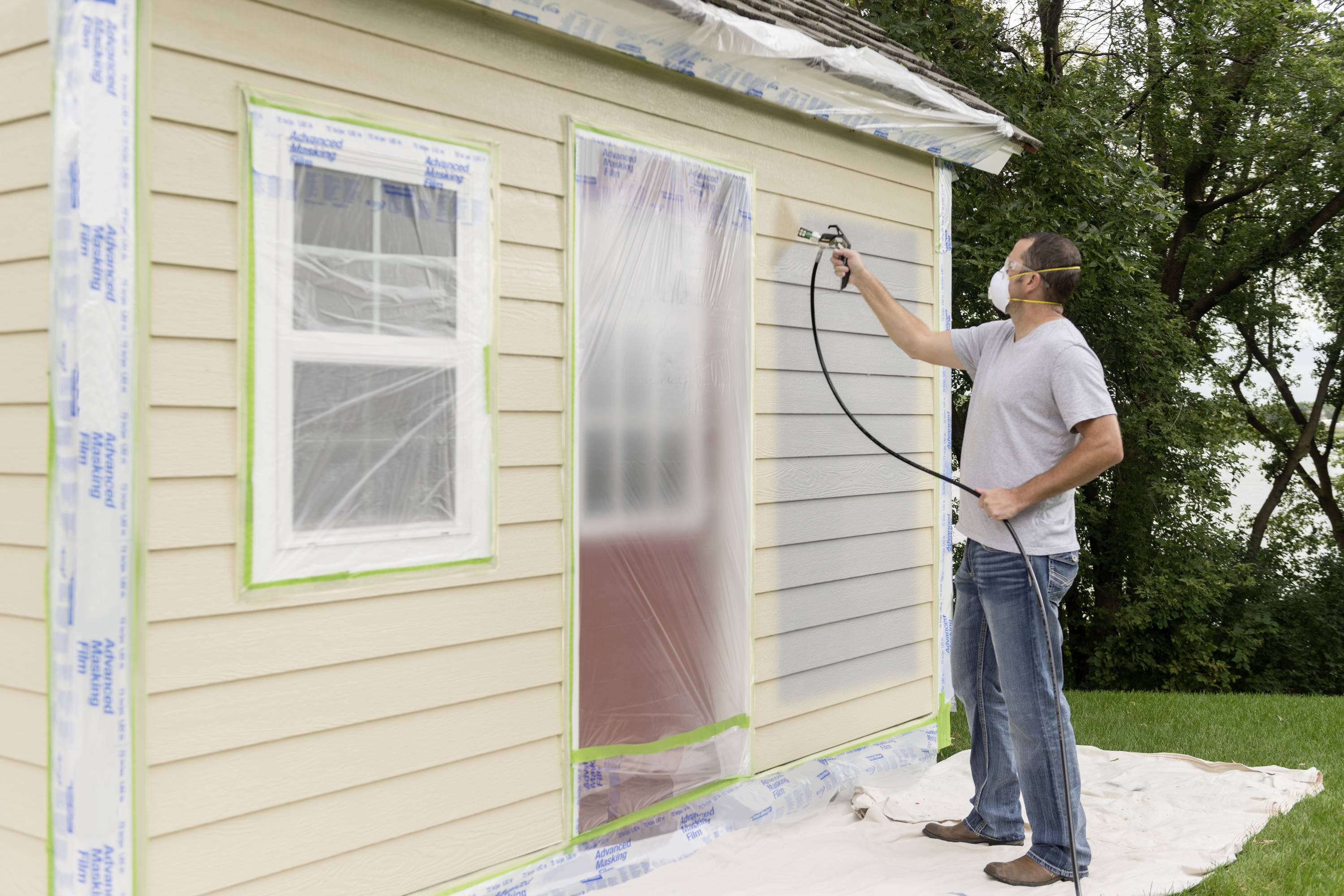 A man is spraying paint on the side wall of a house. Windows and door are covered with plastic sheeting. He is wearing a protective mask.