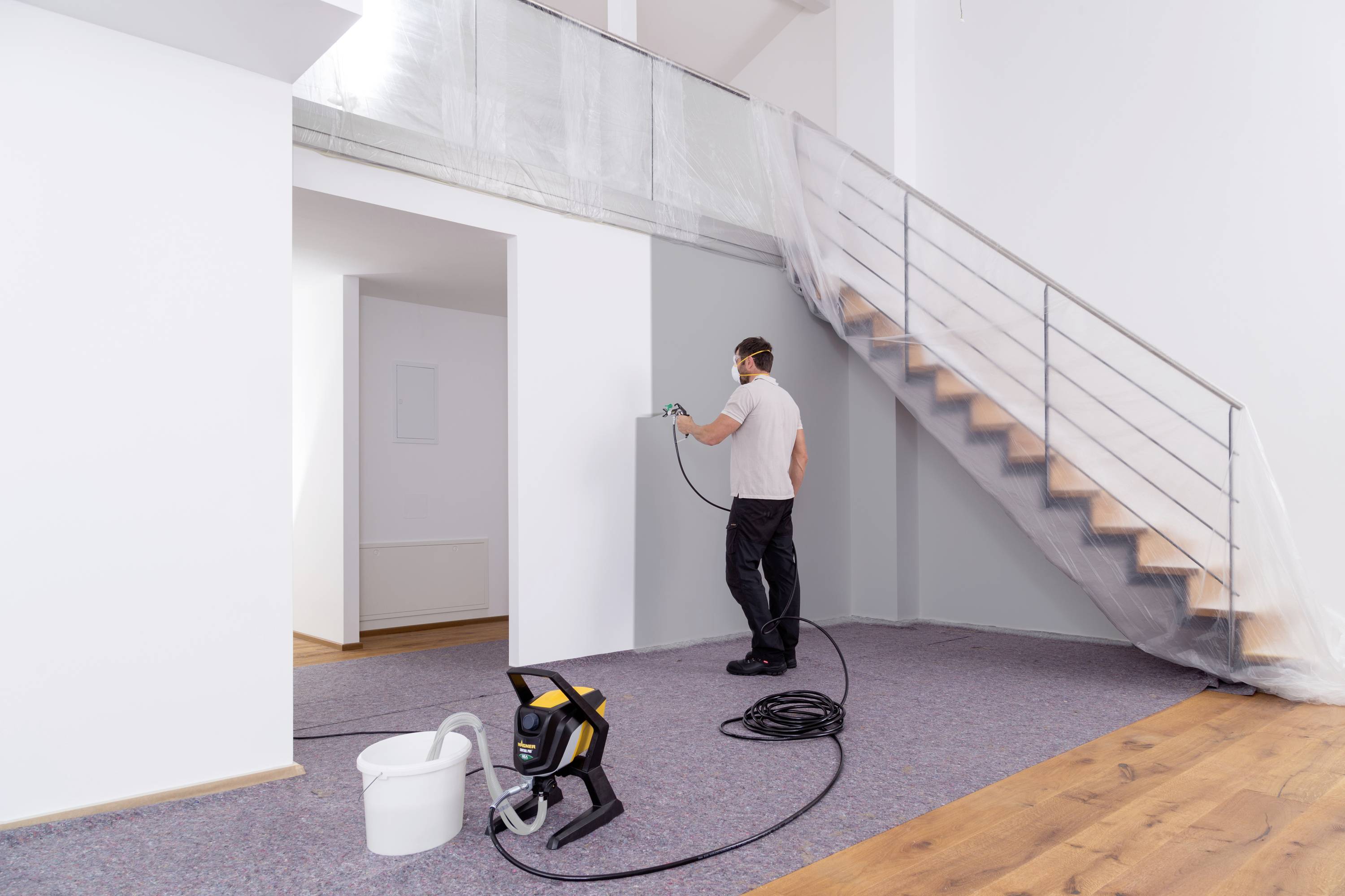 A man is spraying a wall with a spray gun in a modern, white room with a staircase. A paint bucket is positioned in the foreground.