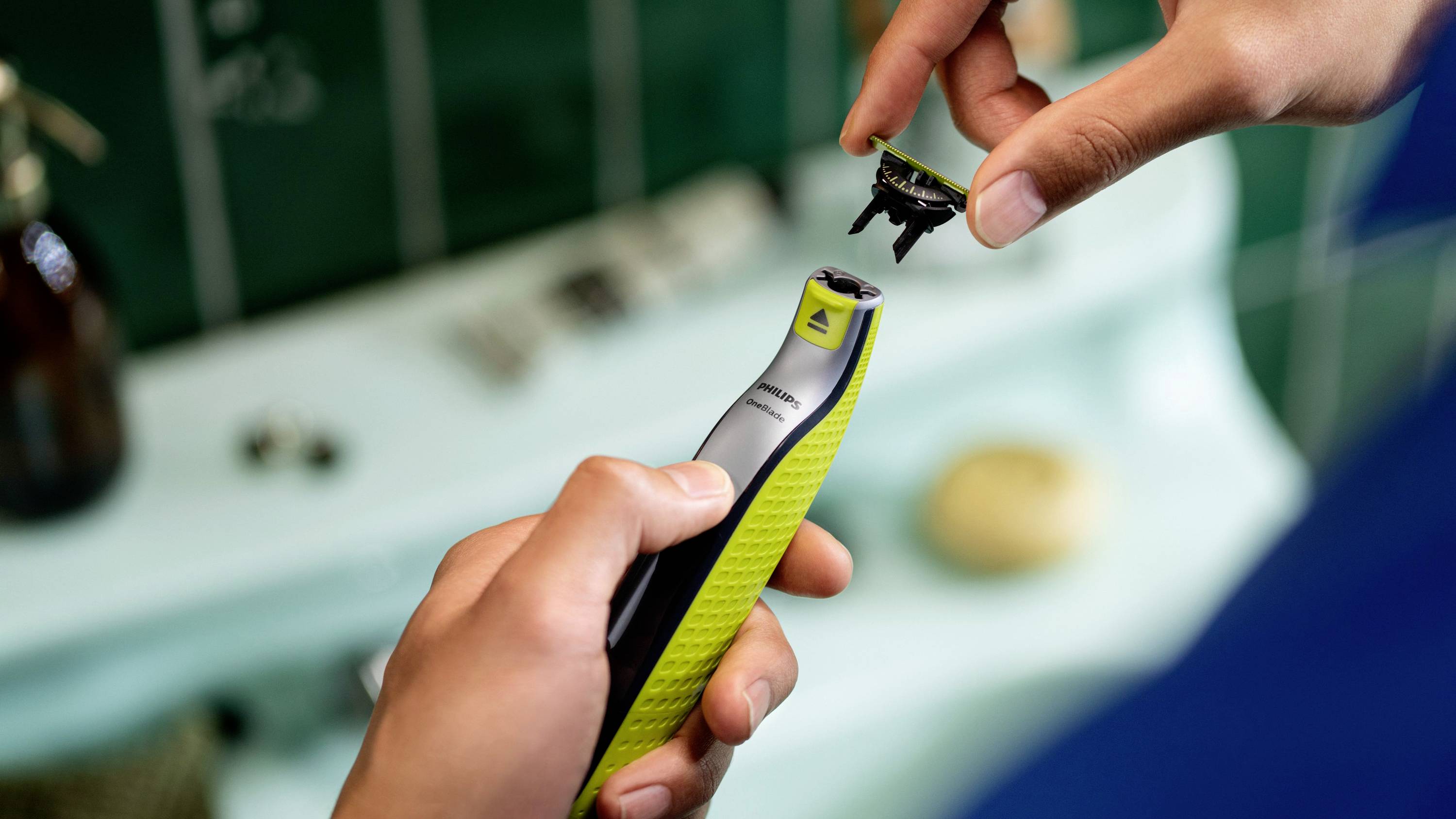 A person is holding an electric razor and changing the razor head. The background shows blurred bathroom accessories.