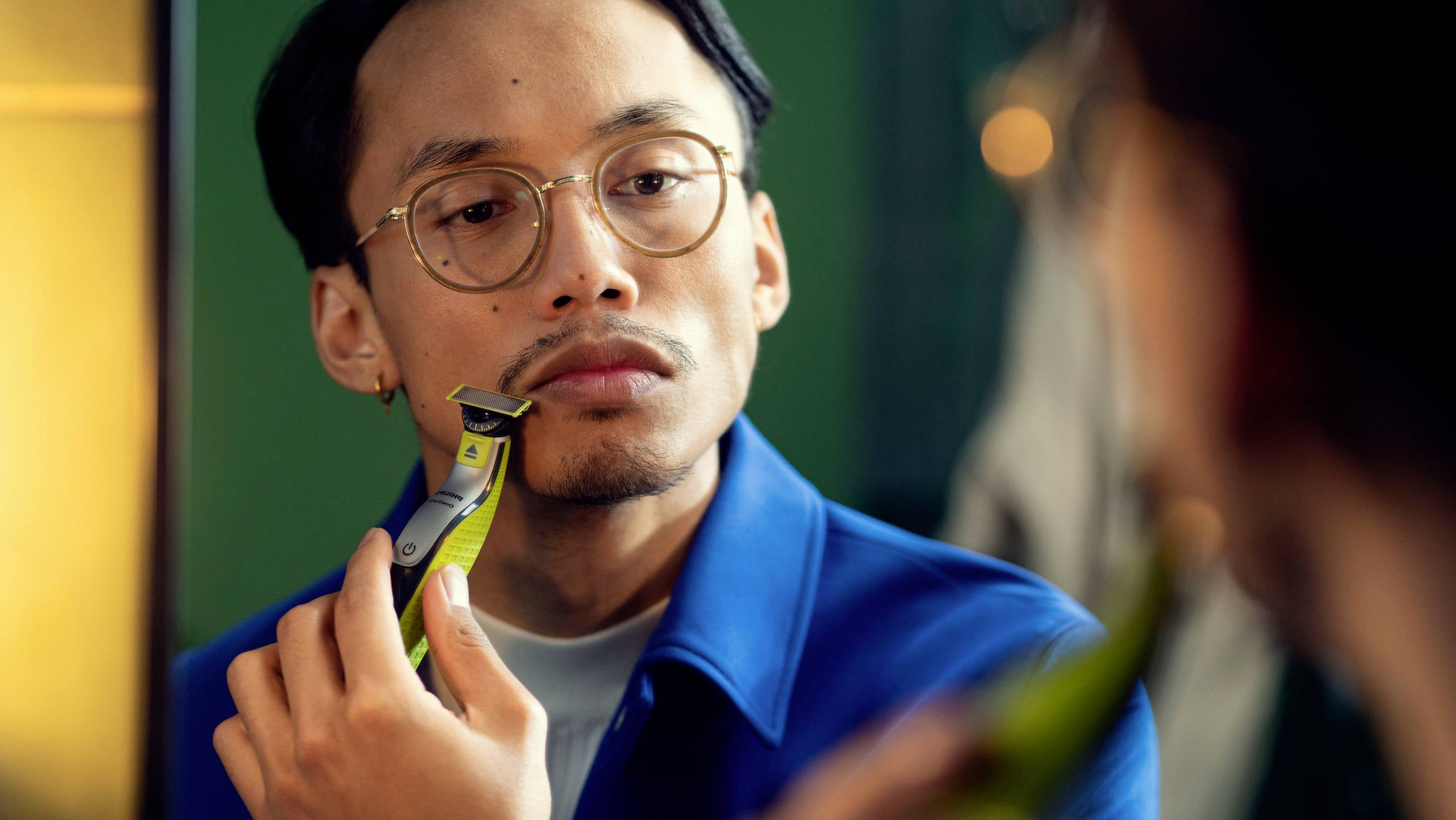 A person wearing glasses is shaving in front of a mirror with an electric razor. They are focusing on their chin.