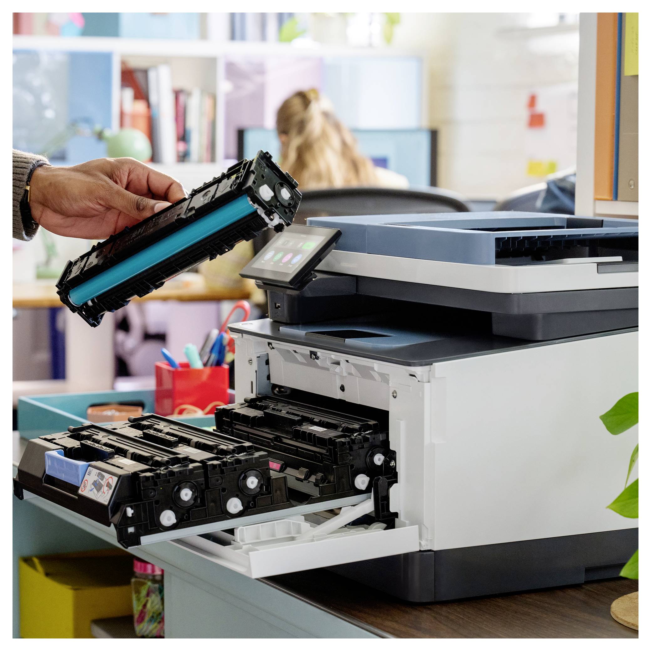 Hand replacing a printer toner cartridge beside an office multifunction printer, with other cartridges in the open tray.
