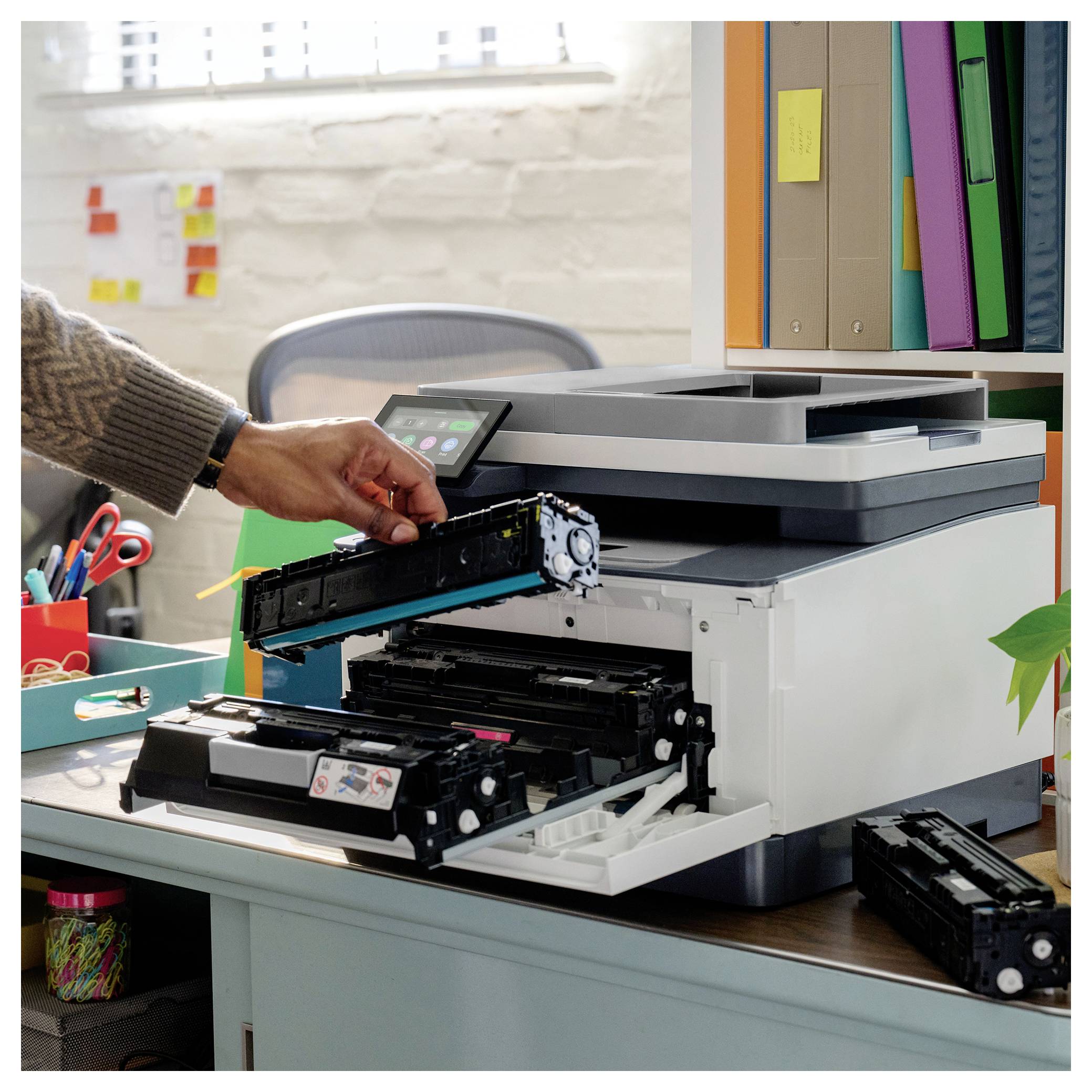 Hand removing a toner cartridge from an open office printer on a desk, with another cartridge nearby and office supplies in the background.