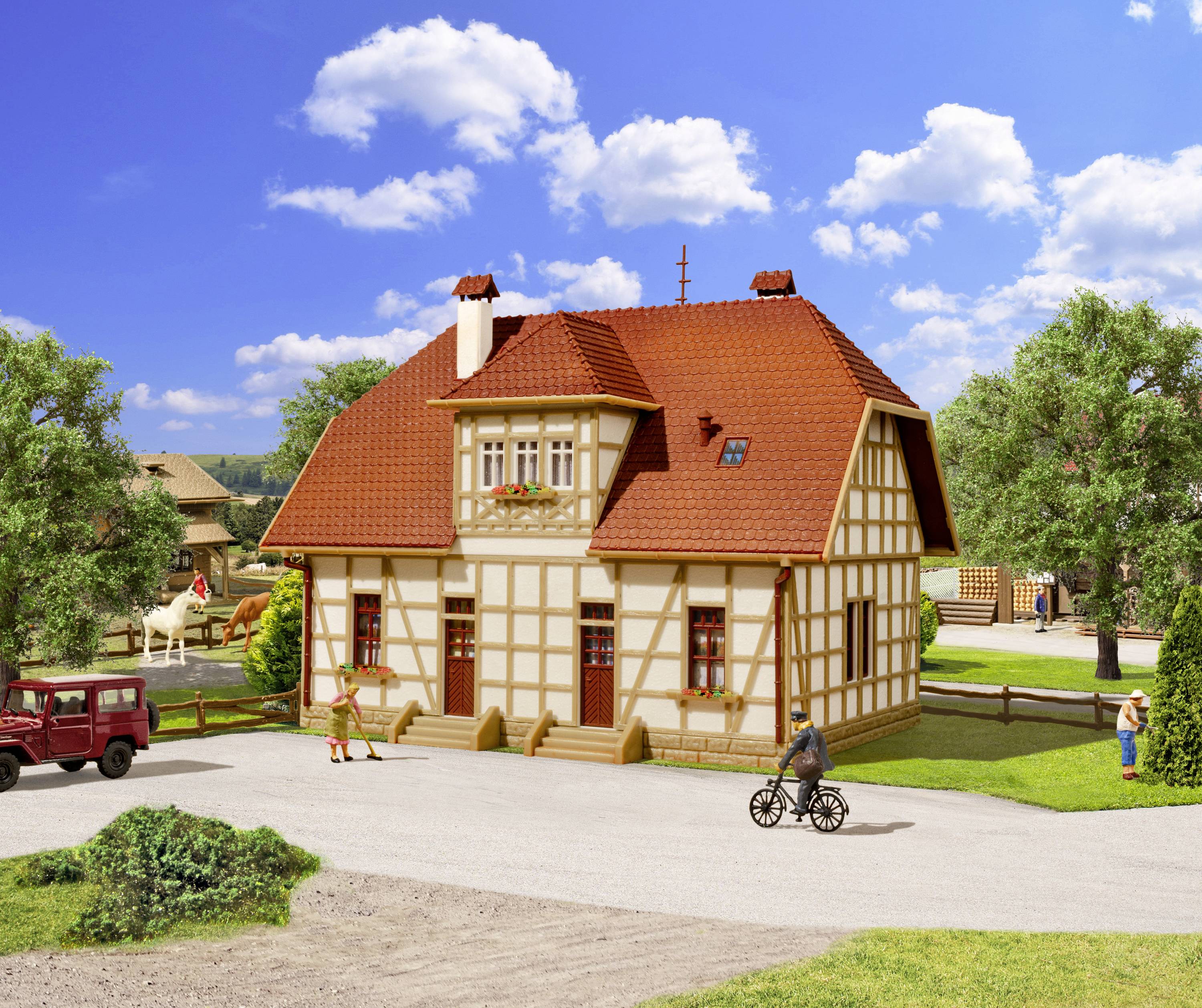 A half-timbered house with a red roof stands in a rural setting. People and a cyclist can be seen on the path in front of it.