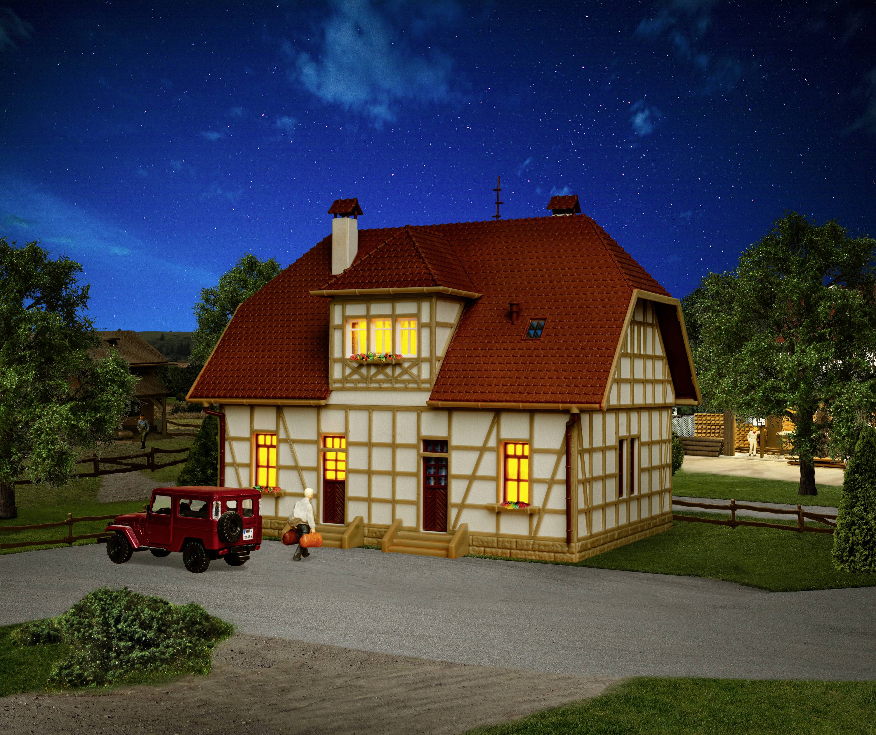A traditional half-timbered house with glowing windows at night, surrounded by trees. A red car is parked in front of it.