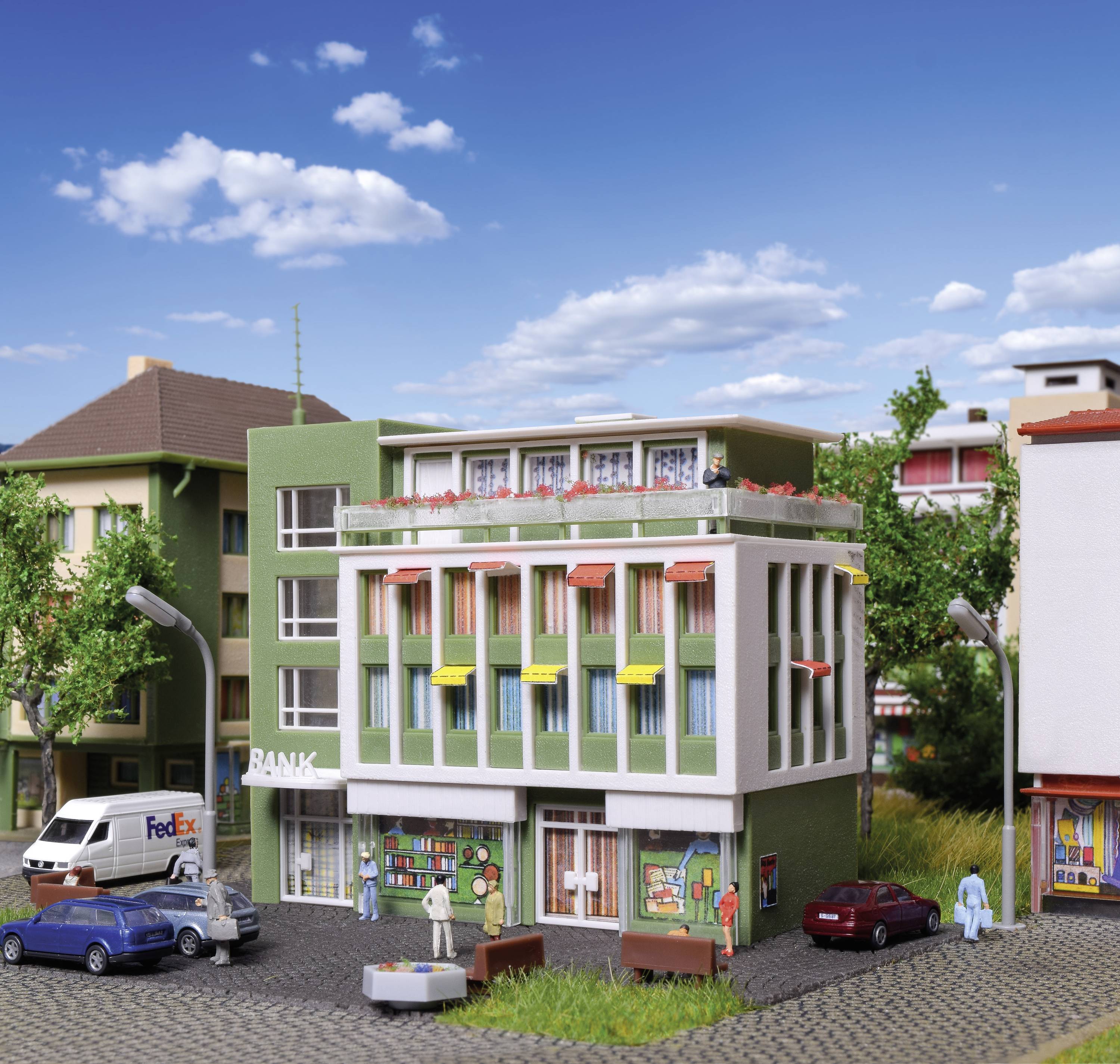 Multi-storey model building with shops on the ground floor and flats above, surrounded by model vehicles and trees.