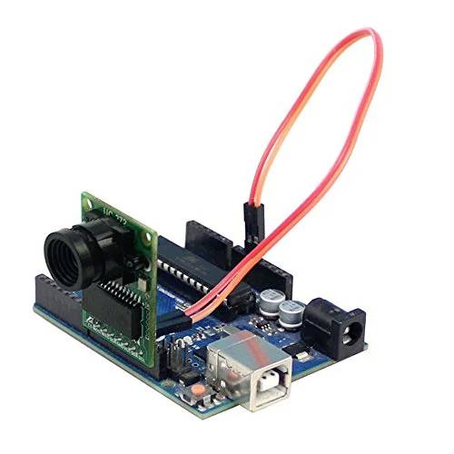 An Arduino board with an attached camera module. Red cables connect the components, which are lying on a white surface.