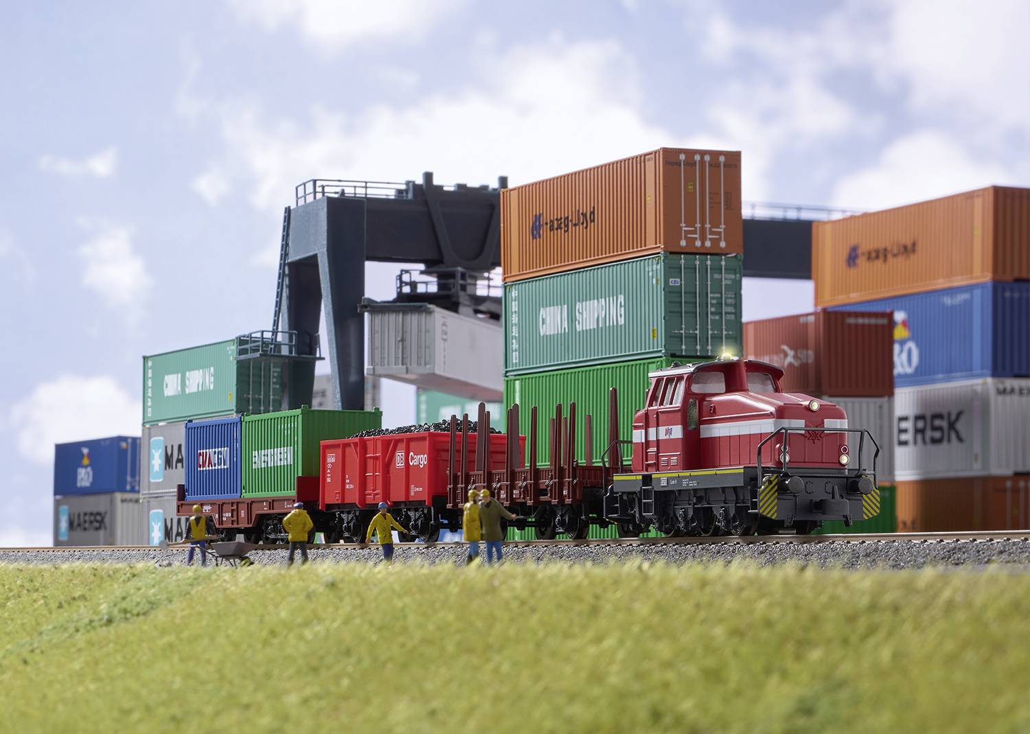 A model train with container wagons is travelling through a model container port. Several figures are interacting with the environment.