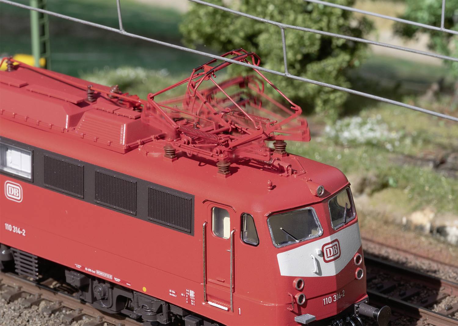 A red model train with a DB logo travels through green countryside under electrical overhead lines on a miniature railway layout.