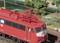 A red model train with a DB logo travels through green countryside under electrical overhead lines on a miniature railway layout.