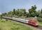 A red train travels through a green, wooded landscape. The sky is blue. This is a model on a model railway layout.