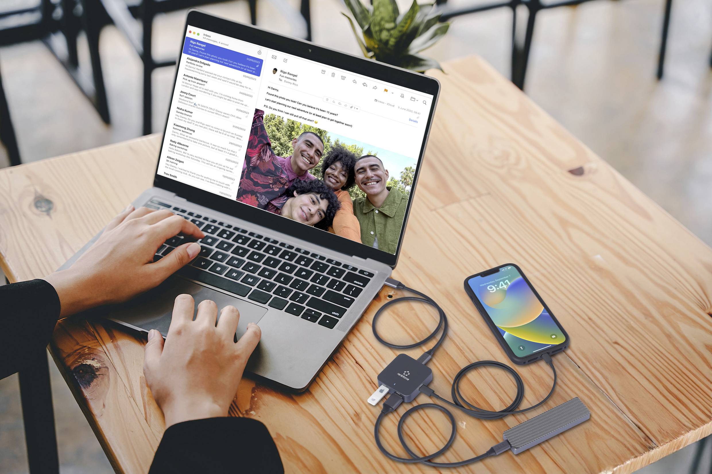 A person is working on a laptop with an open email app, with a smartphone charging nearby; wooden table in the background.