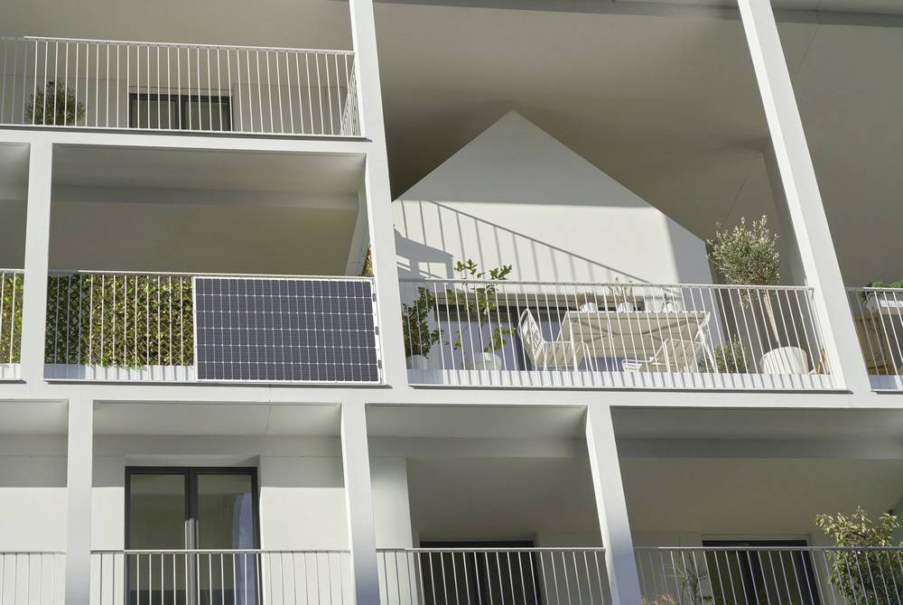 Multi-storey modern building with balconies. One balcony displays solar panels, plants, and garden furniture. Sunlight illuminates the facade.