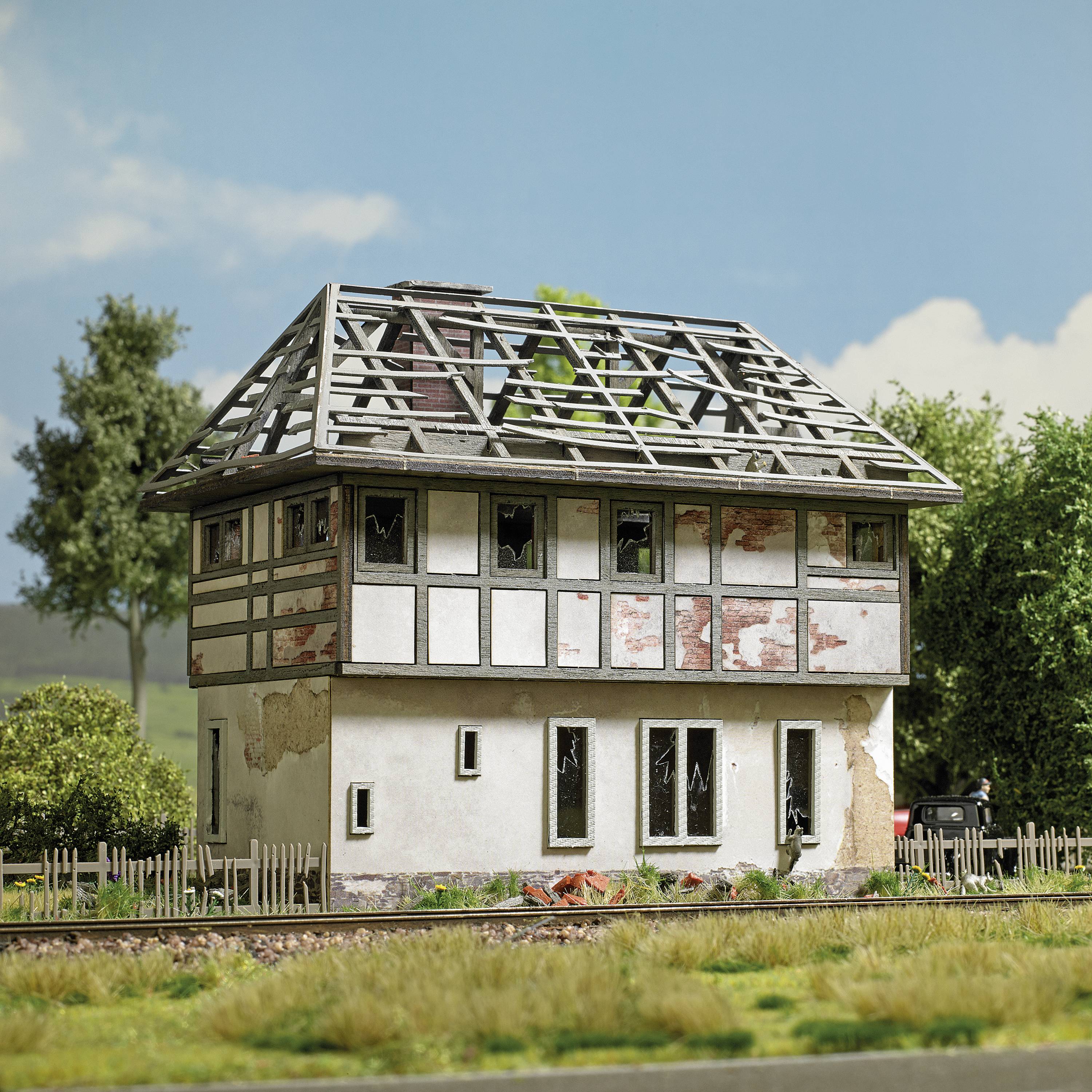 A miniature half-timbered house with a damaged roof stands in a rural setting with trees and a fence in the background.