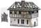 Model of a half-timbered house with visible roof framework, damaged walls and smashed windows. On the left, a bag of accessory parts.