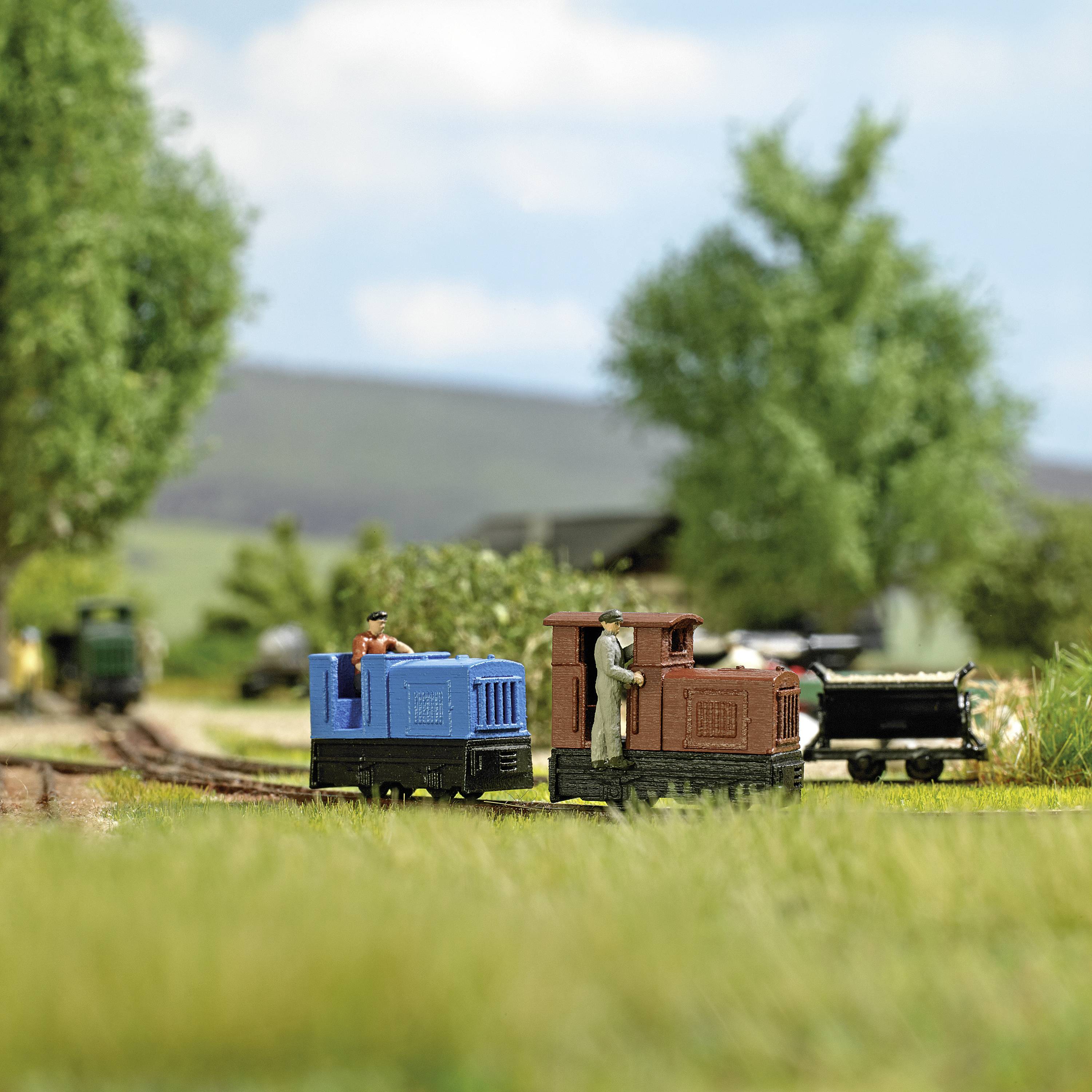 A miniature railway with two locomotives on a track in a rural setting with trees and a hill in the background.