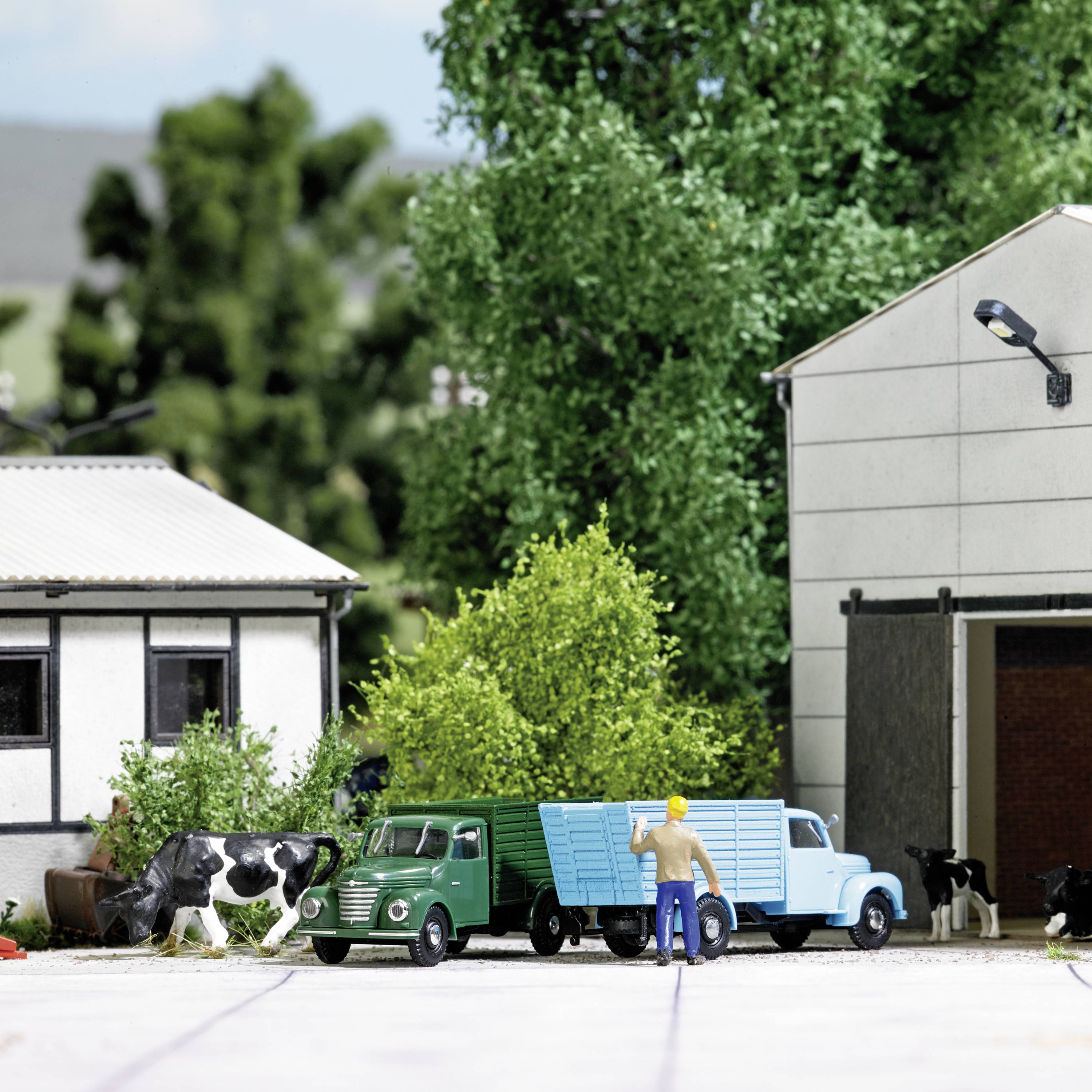 A miniature model shows a green delivery van and a film wardrobe beside buildings and a cow, surrounded by trees and meadow.