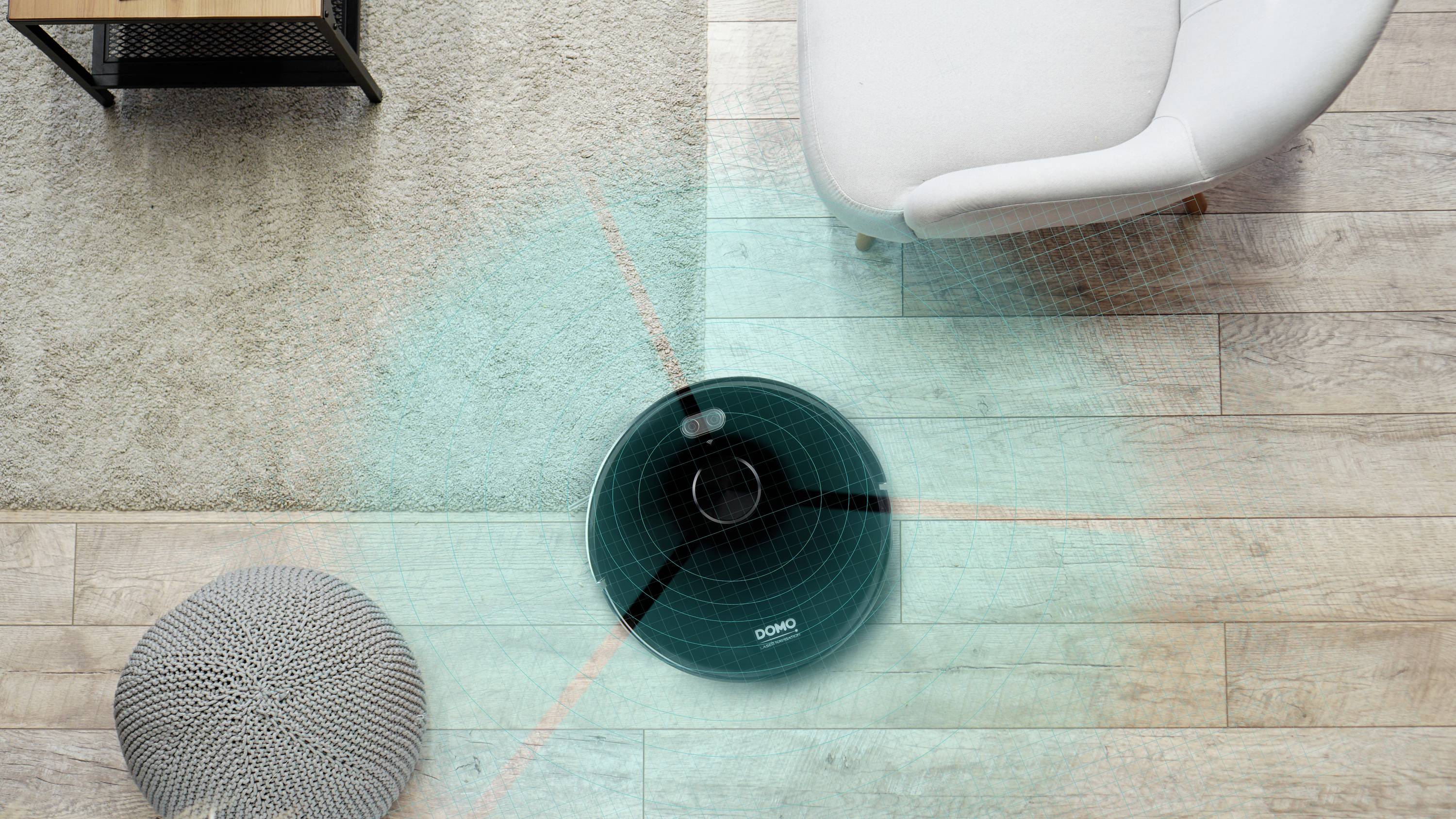 A robotic vacuum cleaner moves across a wooden floor, scanning the area with a blue light beam, a grey pouf to the left, a white armchair to the right.