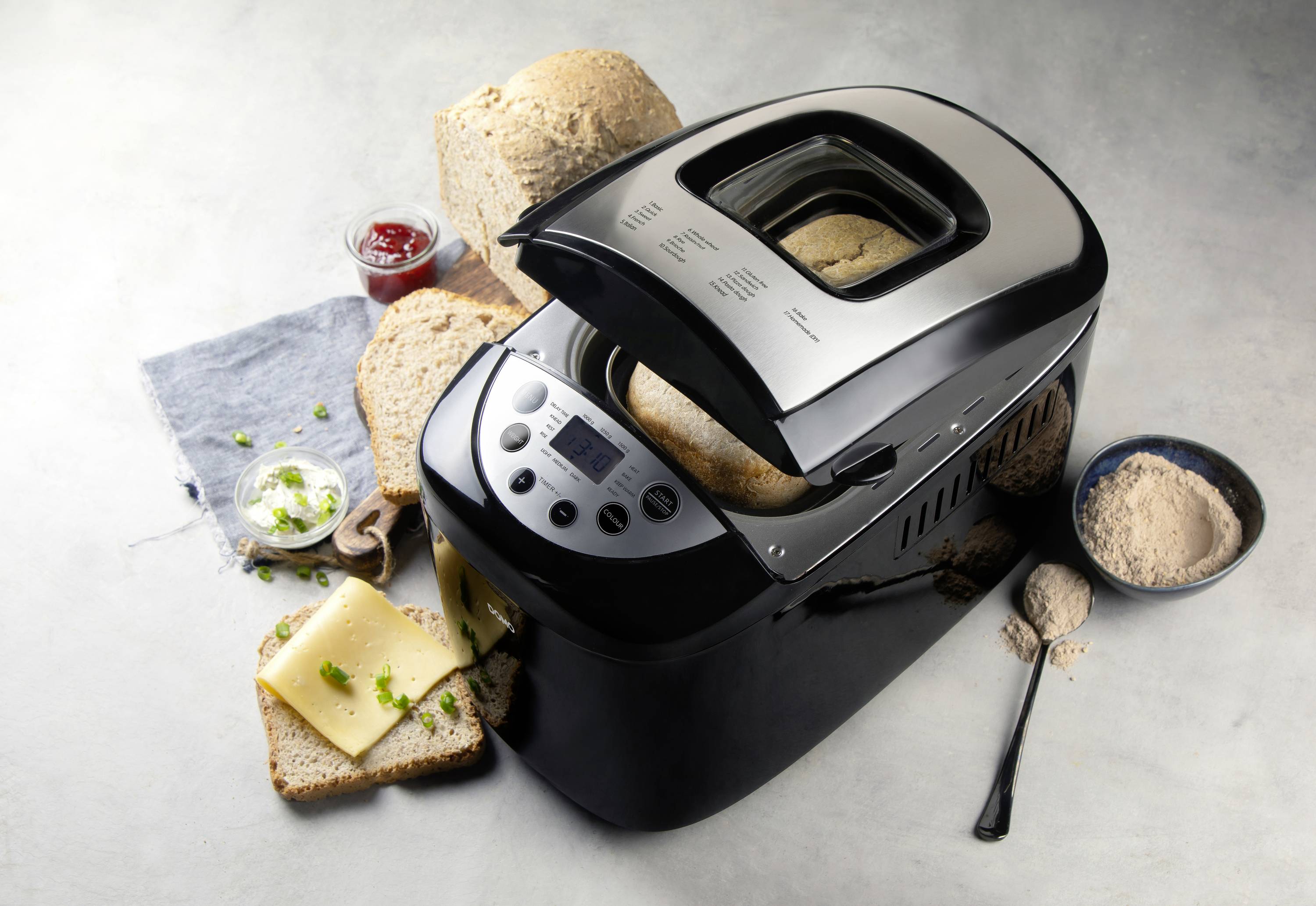 Bread machine with baked bread; beside it sliced bread, cheese and a bowl of flour; ingredients for fresh bread-making.