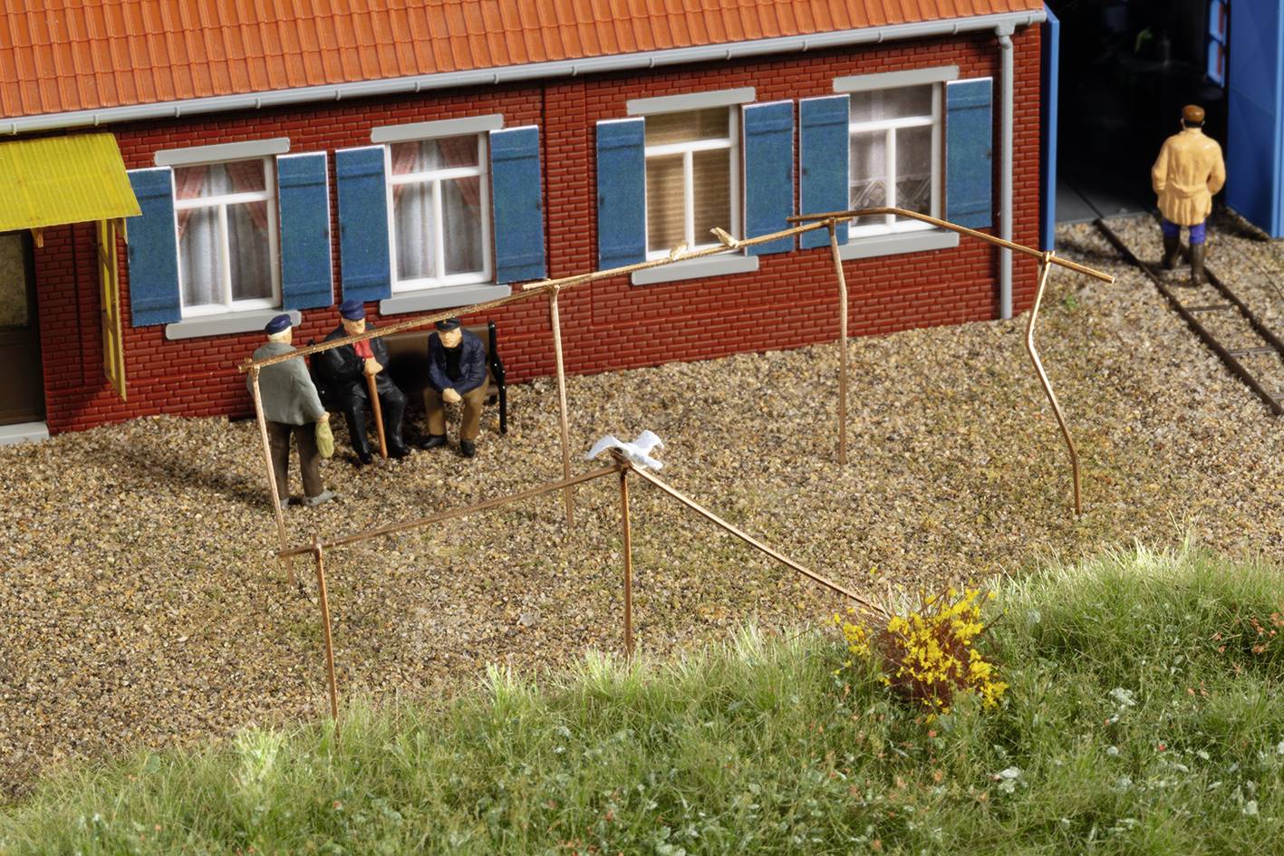 Three people sit and stand in front of a brick house with blue shutters. A bird perches on a low fence.