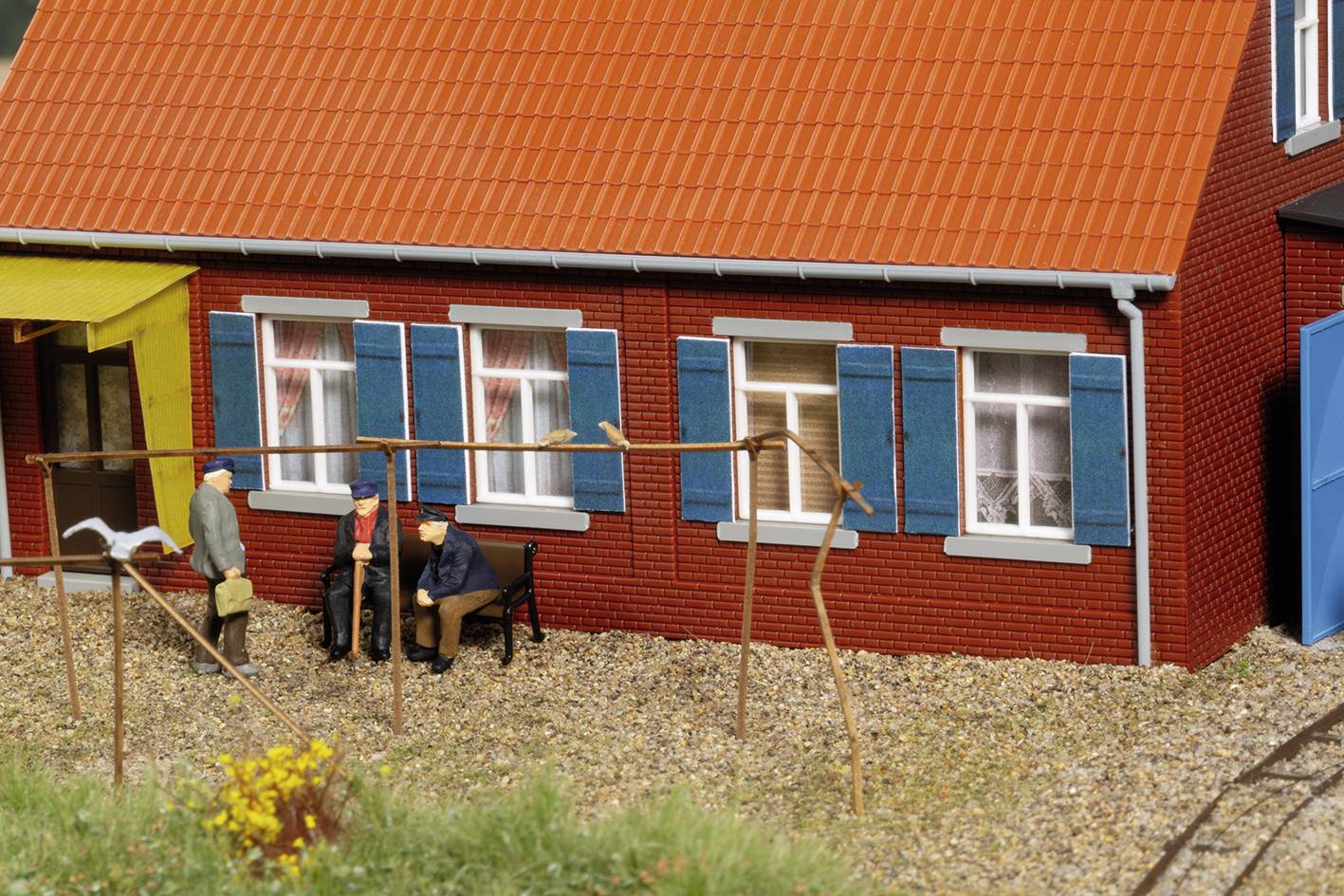 A miniature model of a brick house with a red roof and blue shutters. Three figurines are sitting on a bench in front of it.