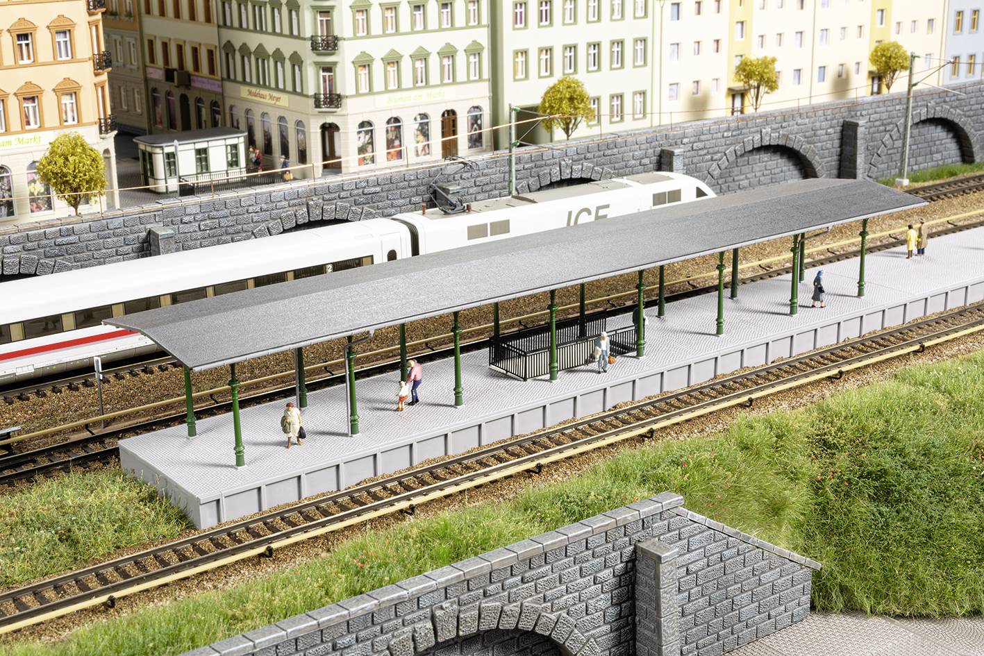 'Model railway scene with high-speed ICE train on tracks. Passengers waiting on the platform. Surrounding urban backdrop with historic buildings.'