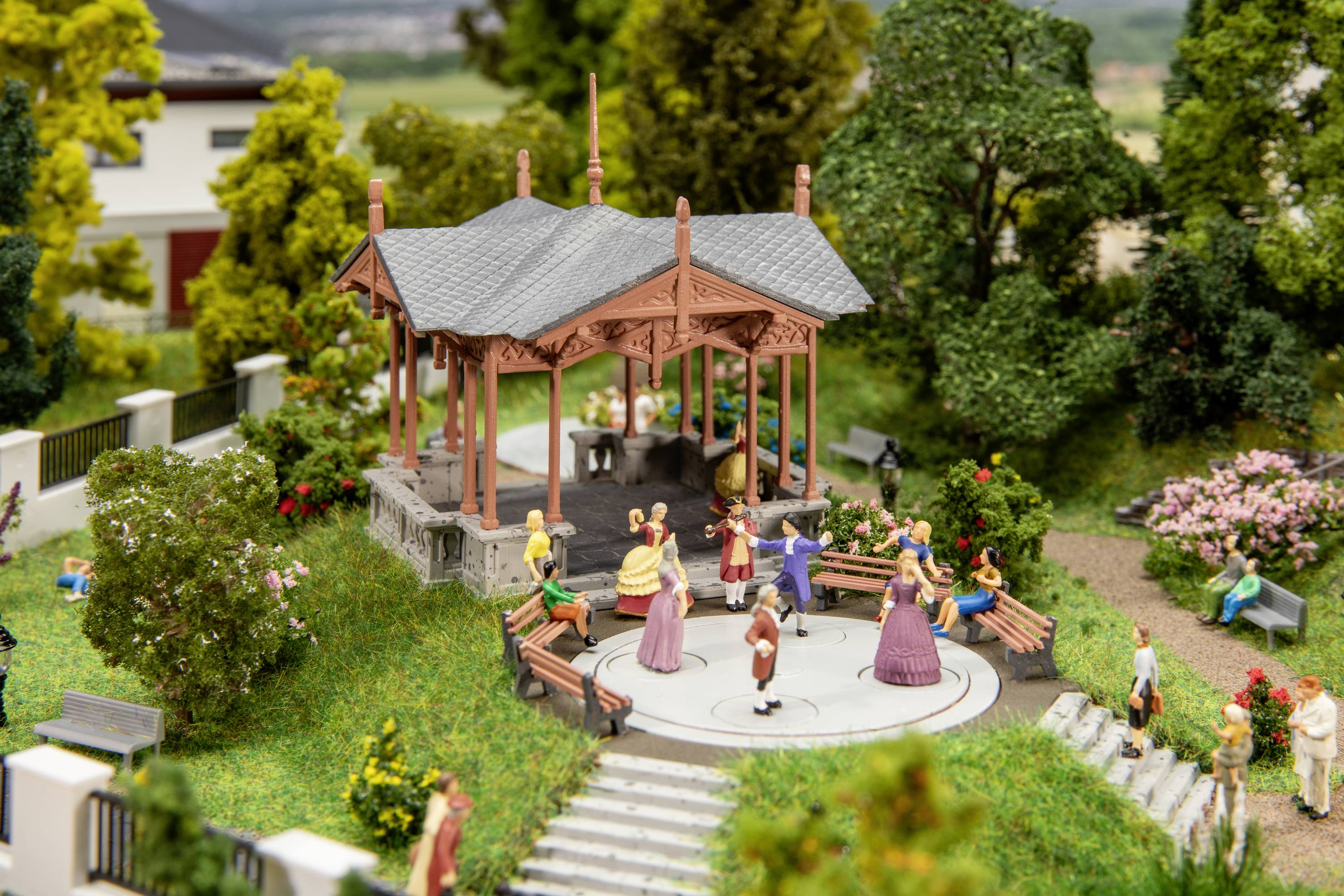 Small figures dance on a stage in a park with trees and benches. It is a miniature scene with a lively atmosphere.