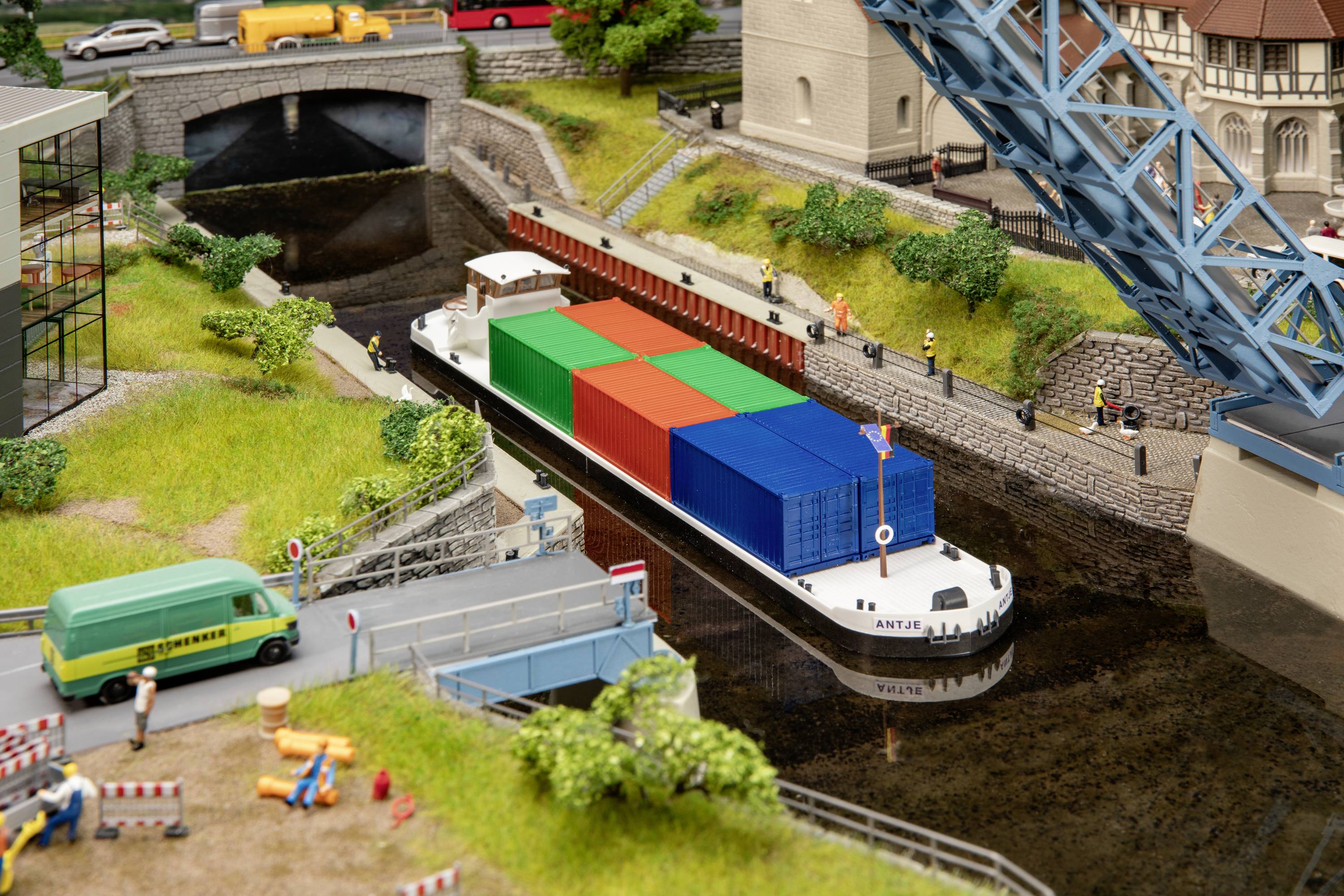 A cargo ship is transporting colourful containers through a canal with a movable bridge. Buildings and vehicles surround the canal.
