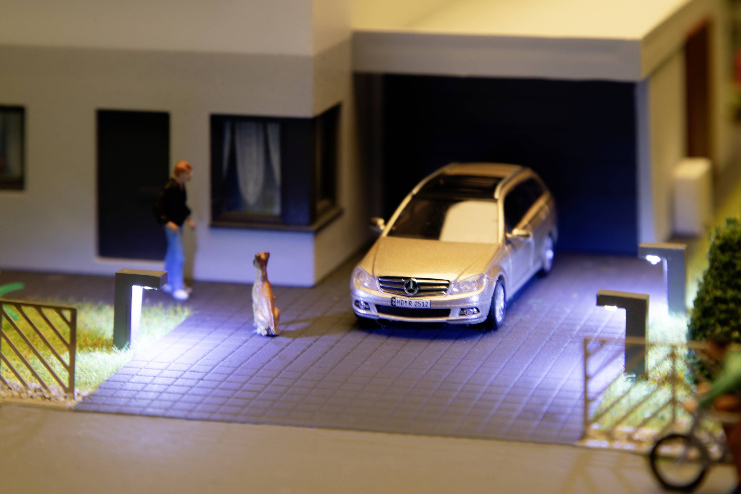 A miniature scene depicts a model car in a lit driveway in front of a modern garage. A figure is standing next to a dog.