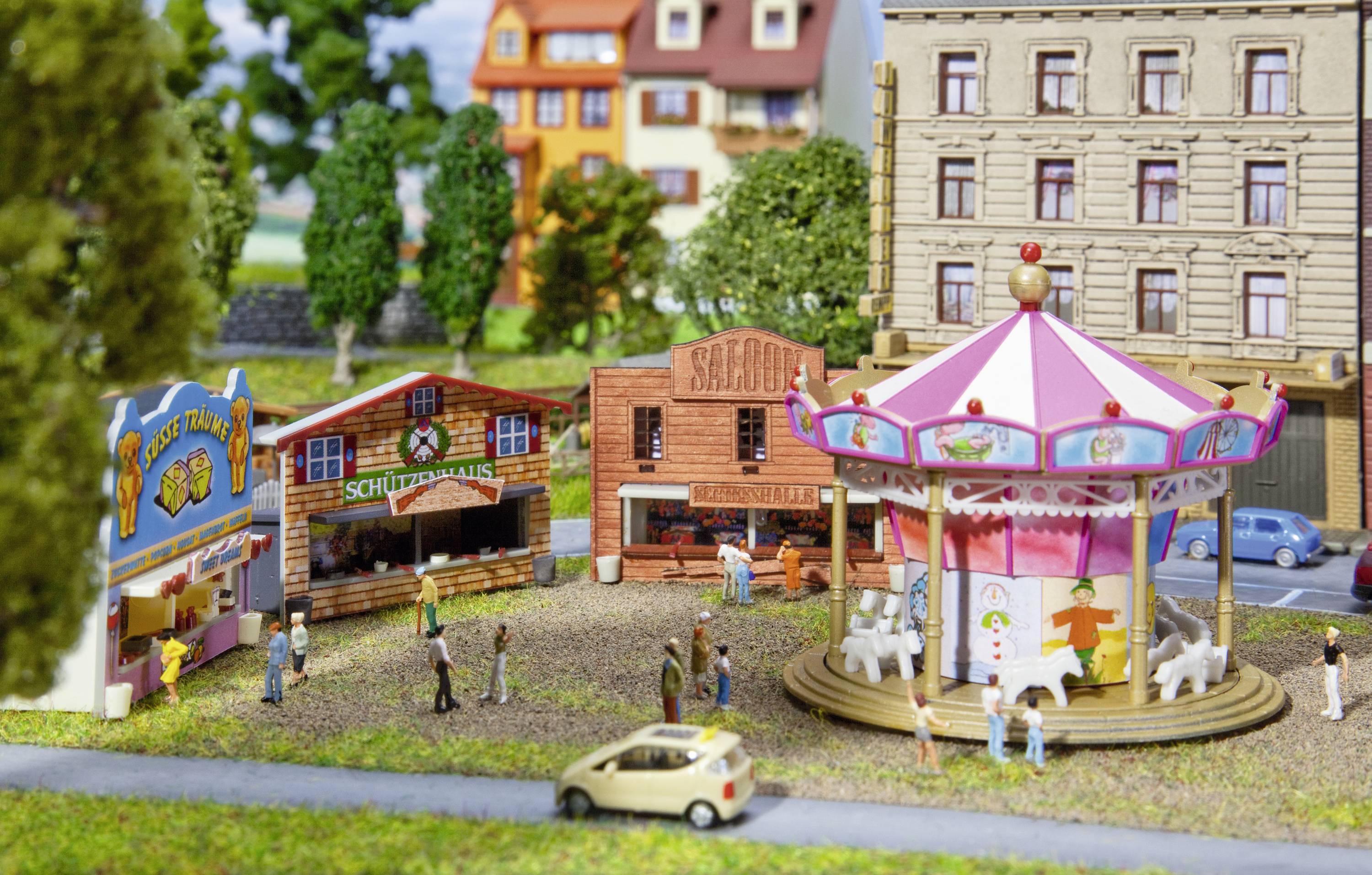A miniature fairground with a carousel, stalls and figures in front of multi-storey buildings. People are walking, some standing at stands.