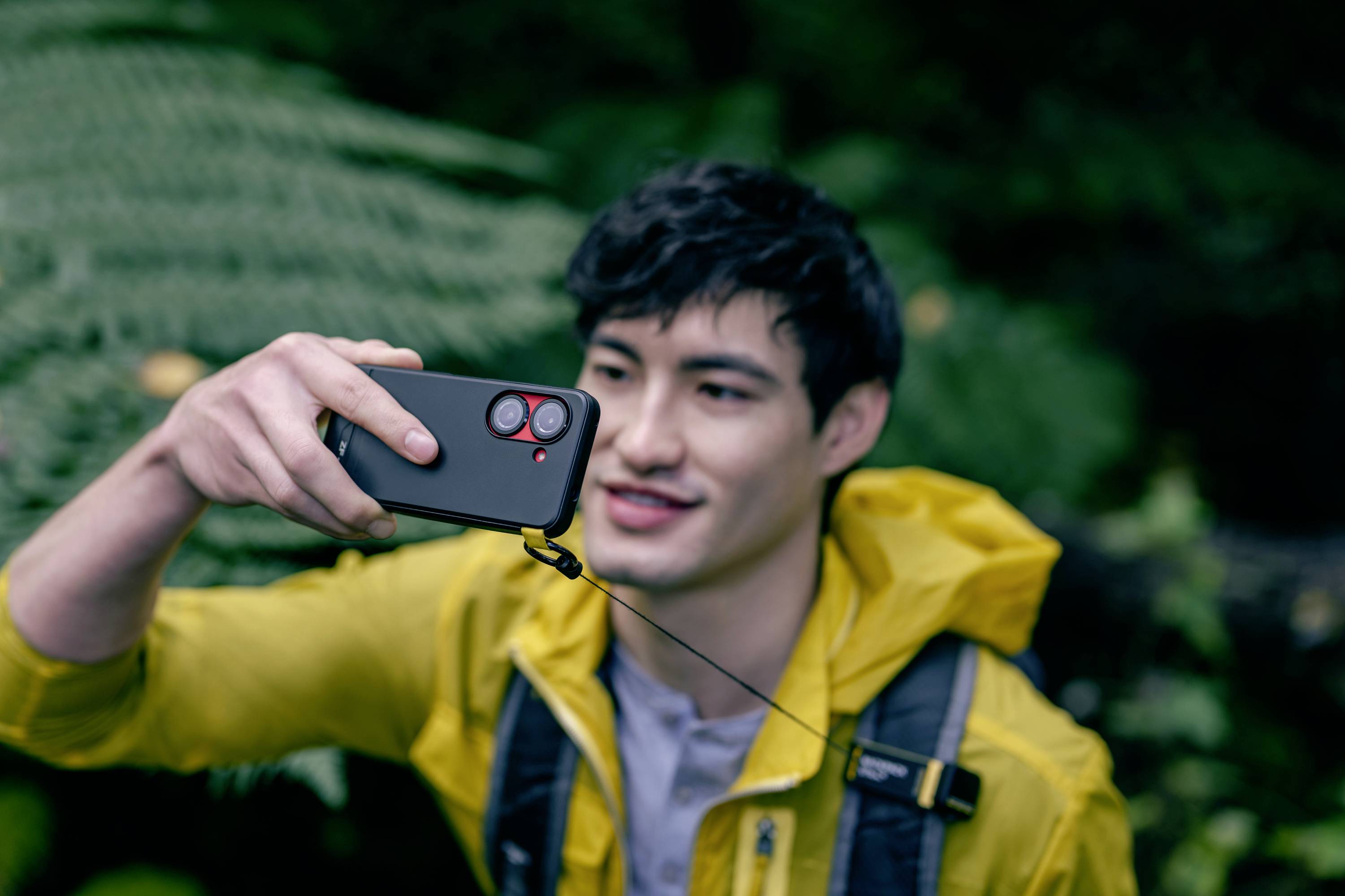 A man in a yellow jacket is holding a smartphone, taking a selfie in a green environment.