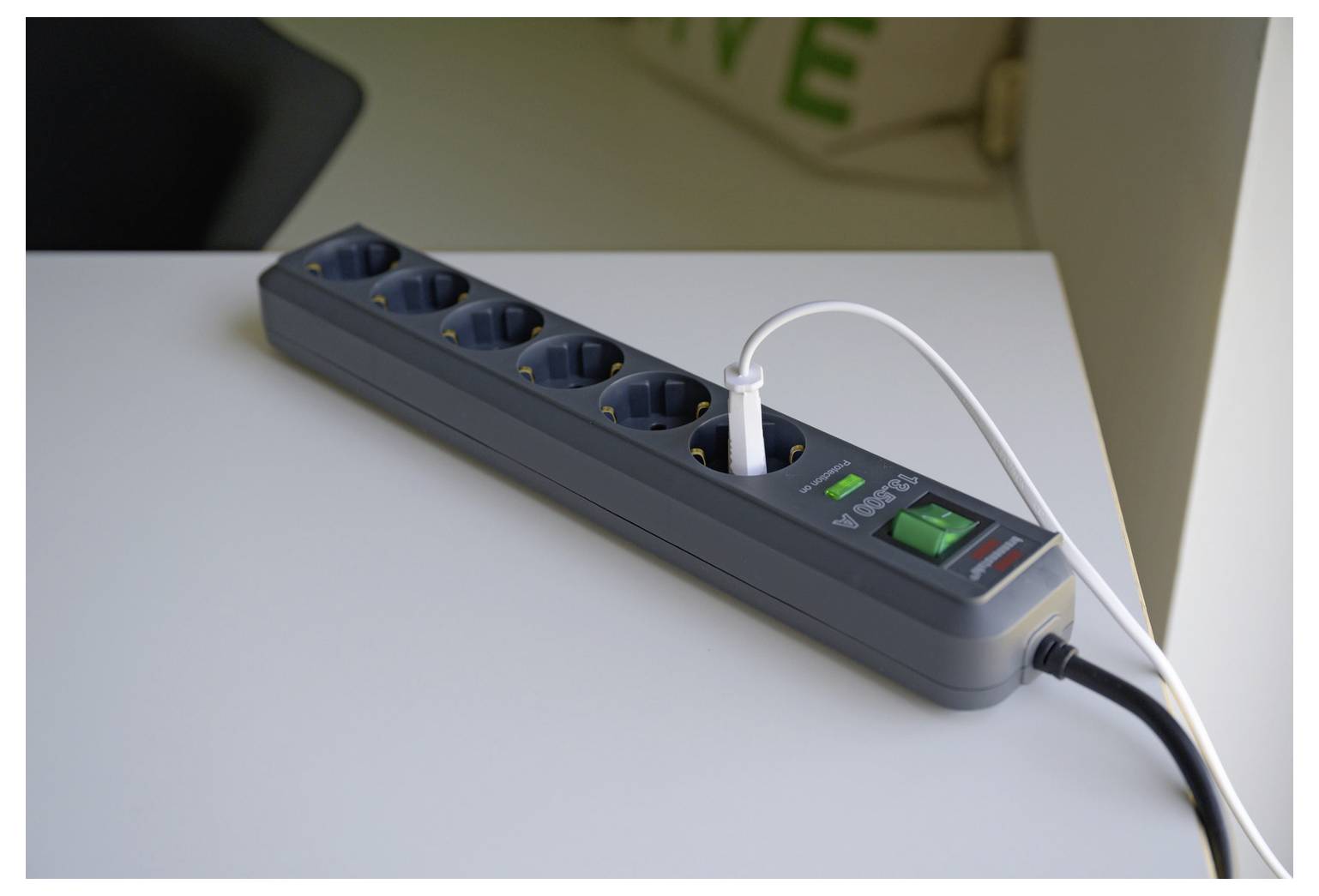 A power strip with several outlets sits on a desk, with a white cable plugged in and a green on/off switch visible.