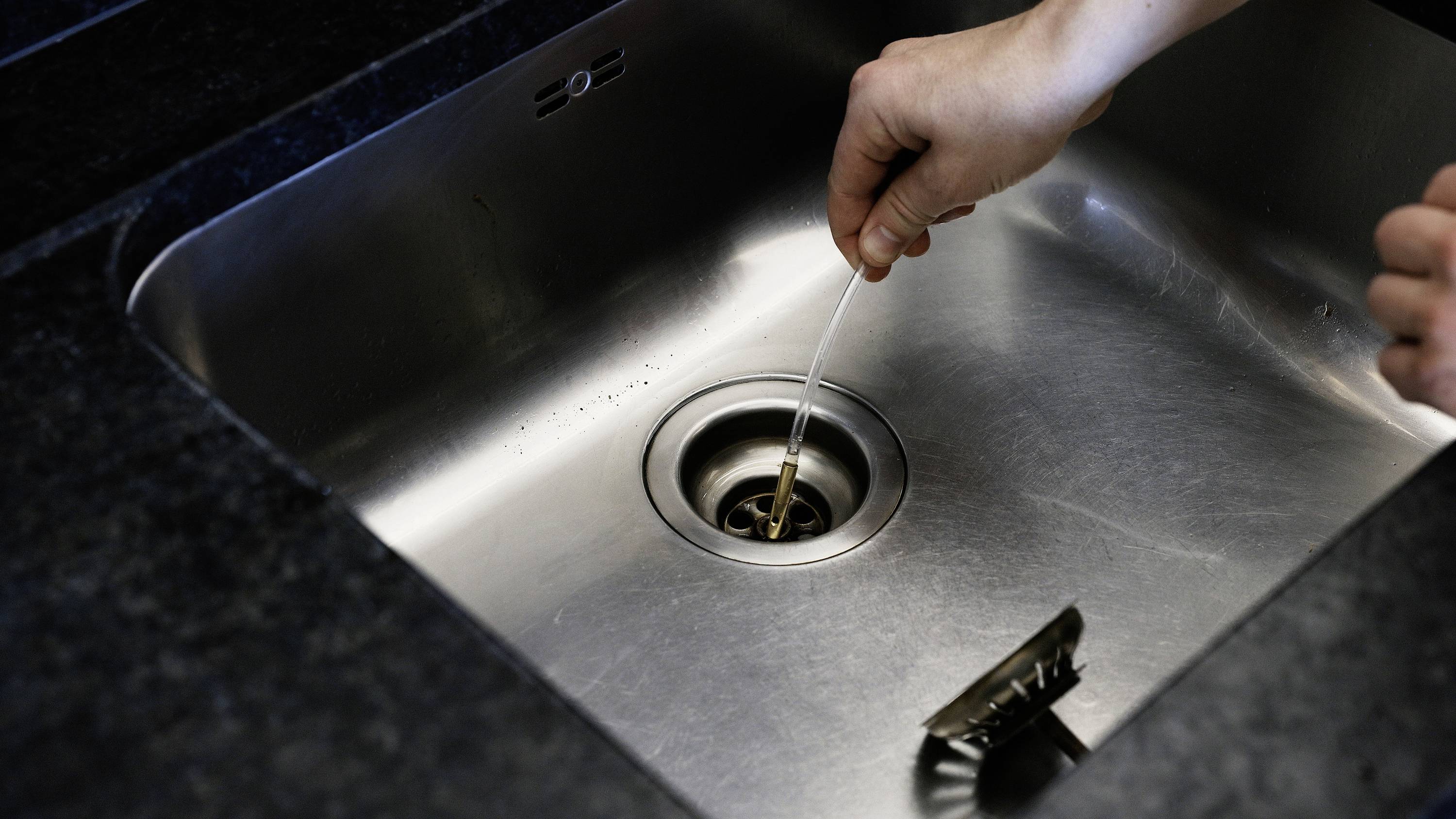 A hand uses a cotton bud to remove something from the plughole of a sink.