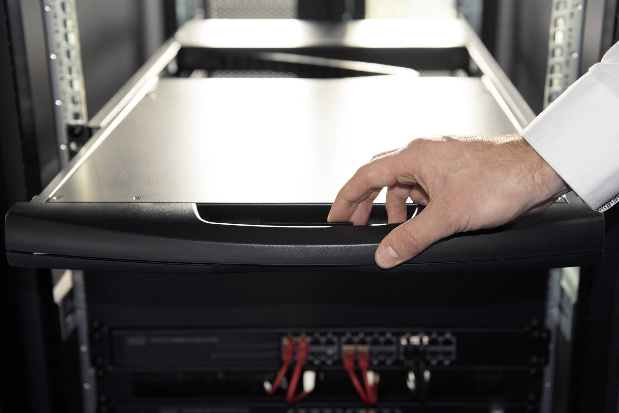 A hand opens a drawer in a server room. Electronic devices and cables are visible in the background.