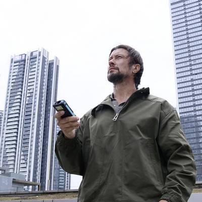 A man outdoors holding a smartphone, with skyscrapers in the background. He is wearing a khaki jacket.