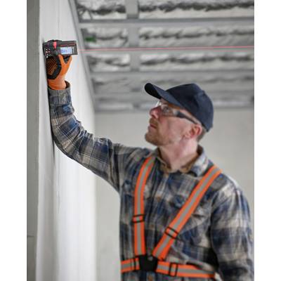 Construction worker in protective clothing measures the wall with a laser distance meter. Ceiling incomplete, revealing metal joists.