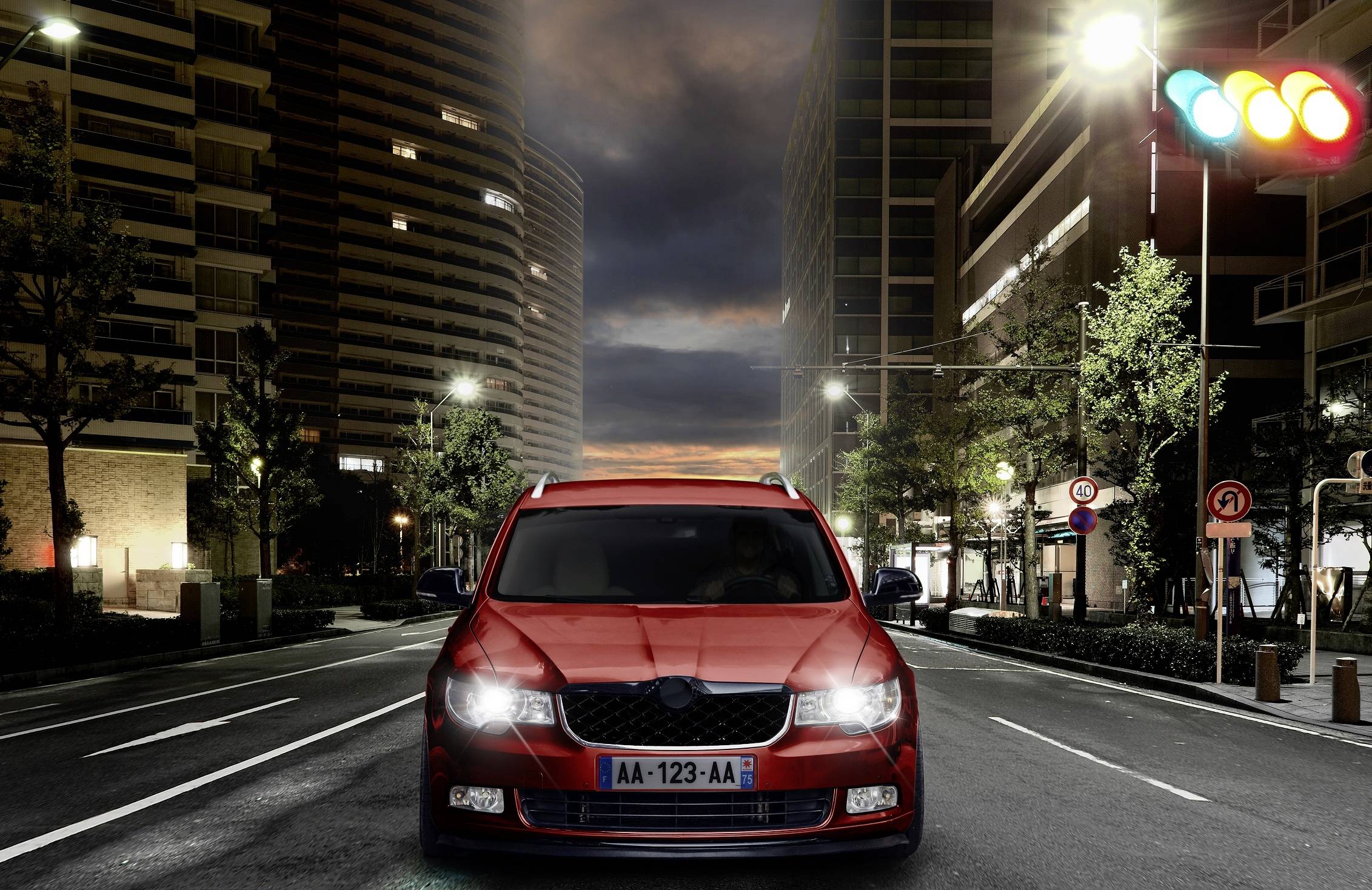 A red car drives through a lit city street at night, stopping at traffic lights with red, yellow, and green lights.