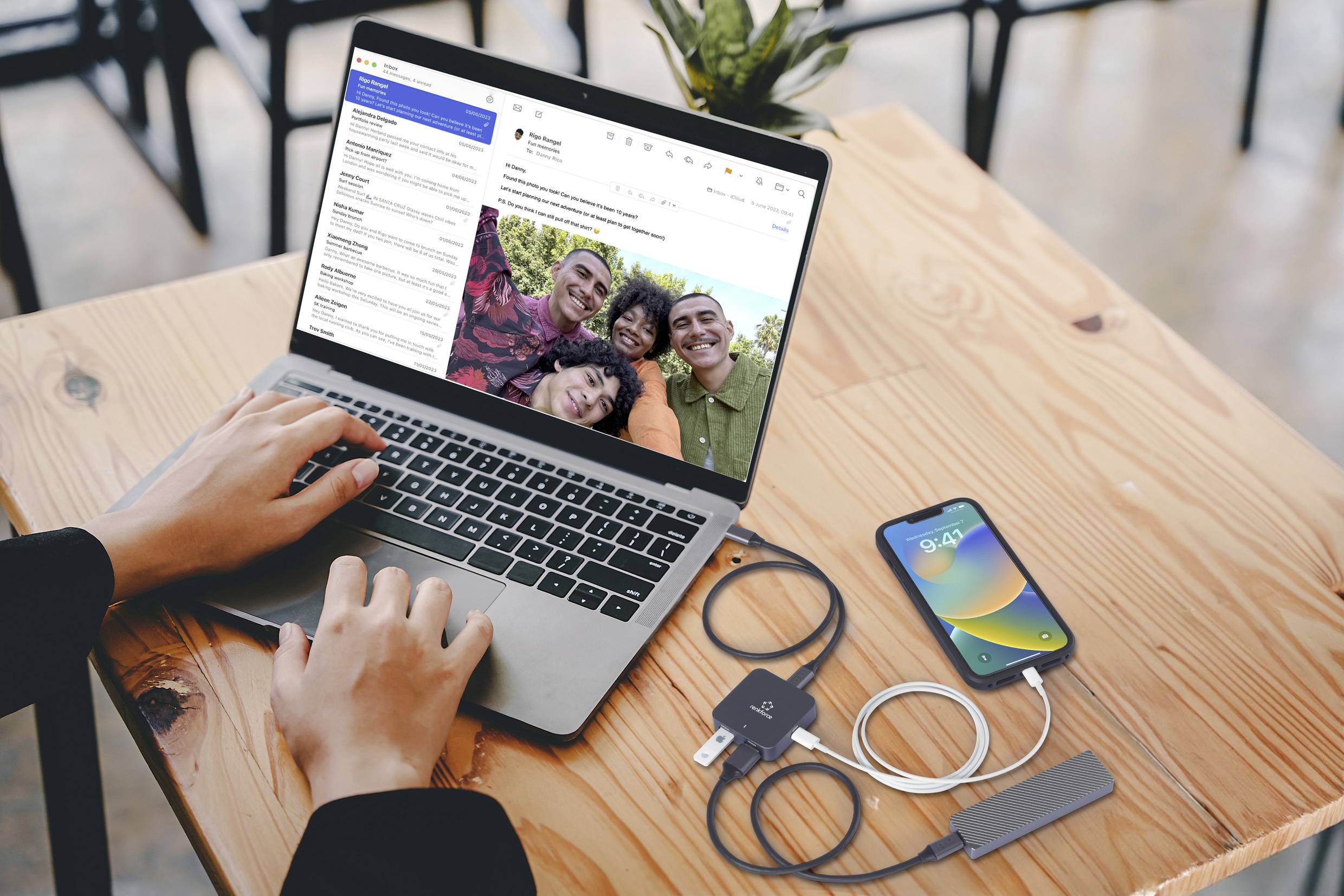 A person is typing on a laptop with an open email inbox, with a smartphone and charging cable, along with other cables, placed beside it on a wooden table.