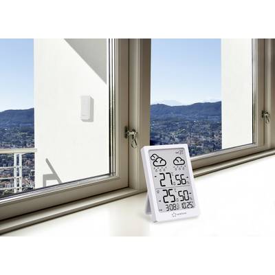 A thermometer displays indoor temperature (27.5°C), outdoor temperature (25.6°C) and air pressure (1012.5 hPa) on its display, positioned in front of a window with a view.