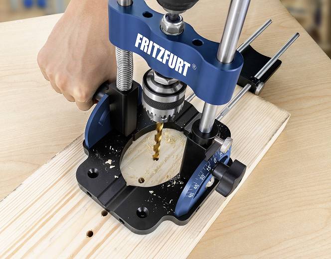 A person is drilling a hole in a wooden board using a blue drill stand. The drill stand is marked with the inscription 'FRITZFURT'.