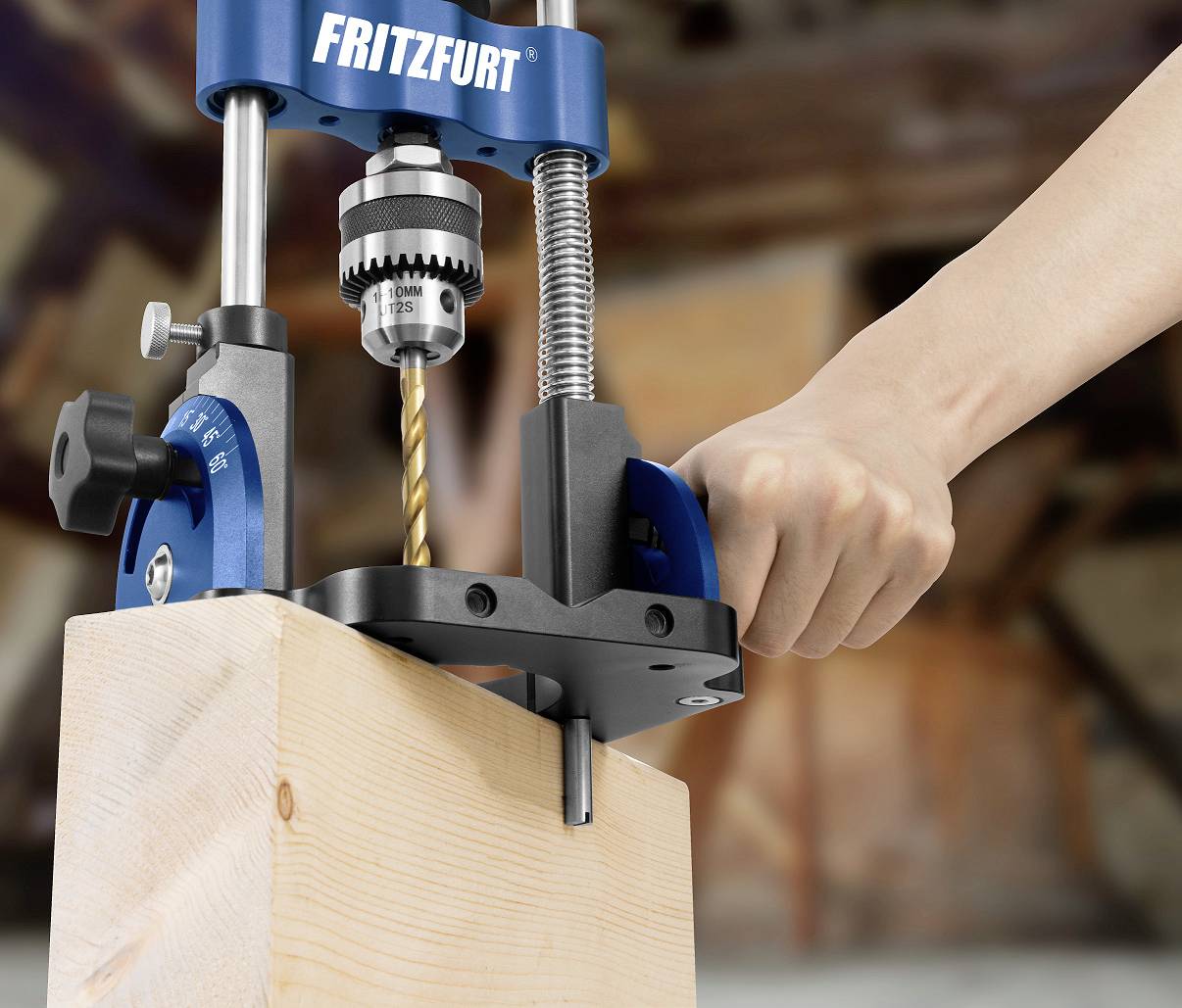 A hand operates a blue pillar drill, boring a hole into a wooden block. The background is out of focus.