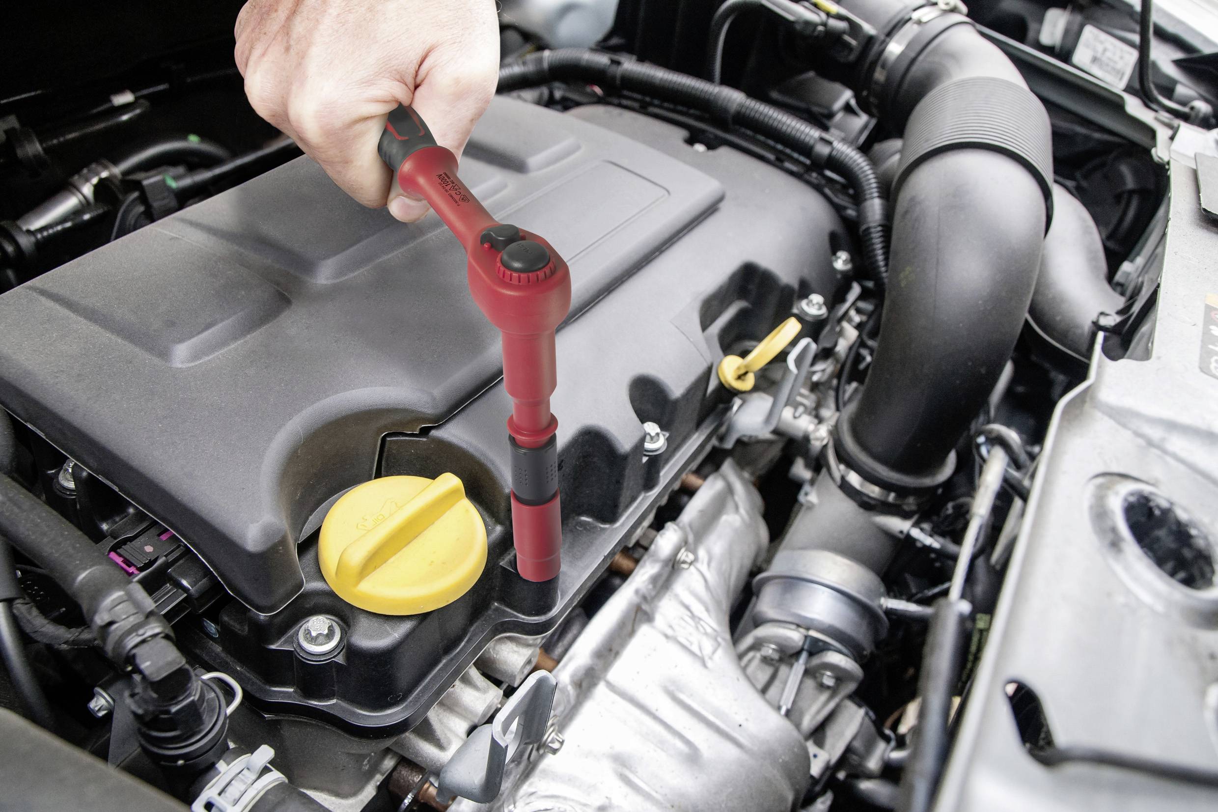 A hand is using a red tool to loosen the spark plug connector in a car engine. Parts of the engine are visible.