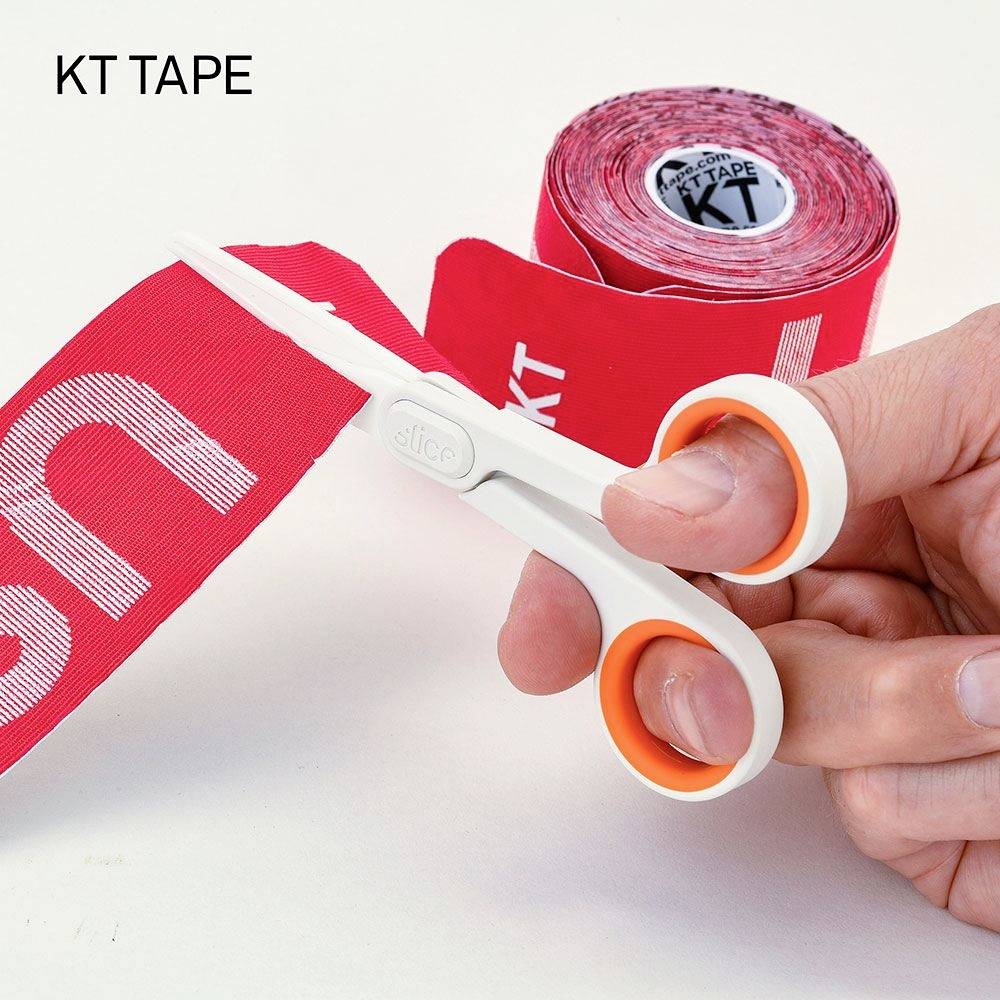 A hand is cutting a piece of red kinesiology tape with a white print from a roll using scissors.