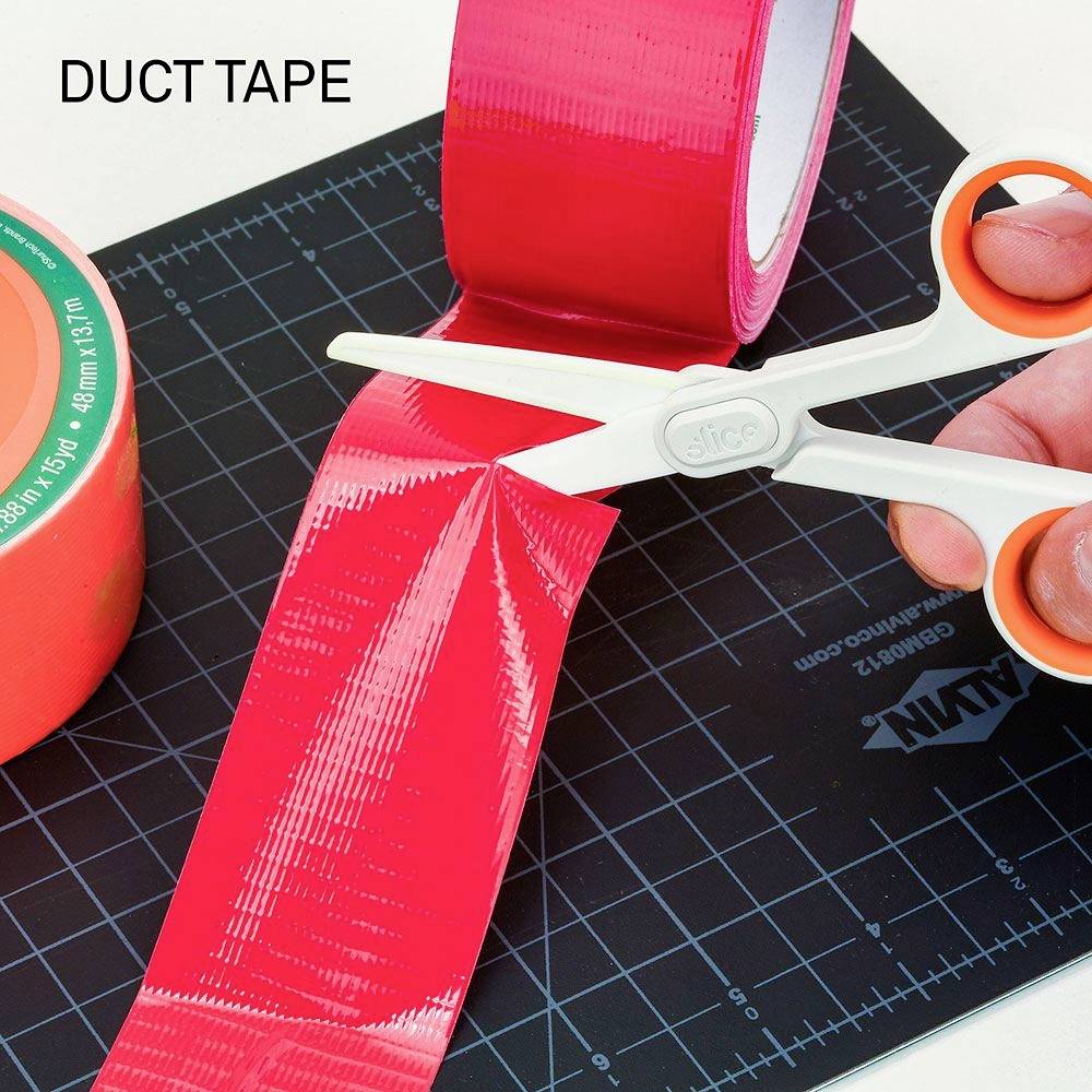 A hand is cutting red adhesive tape with scissors. Next to it, a roll of orange adhesive tape lies on a cutting mat.