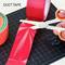 A hand is cutting red adhesive tape with scissors. Next to it, a roll of orange adhesive tape lies on a cutting mat.