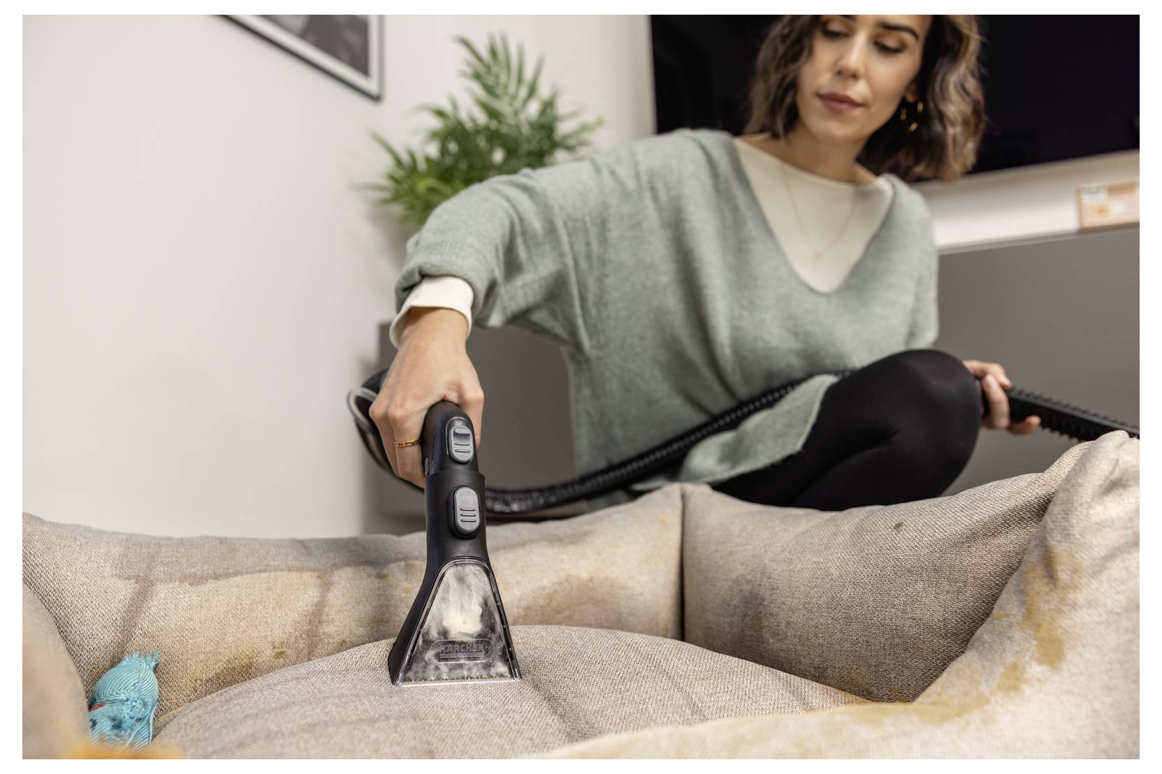 A person uses a handheld vacuum with a brush attachment to clean a fabric sofa. A small plant is visible in the background.