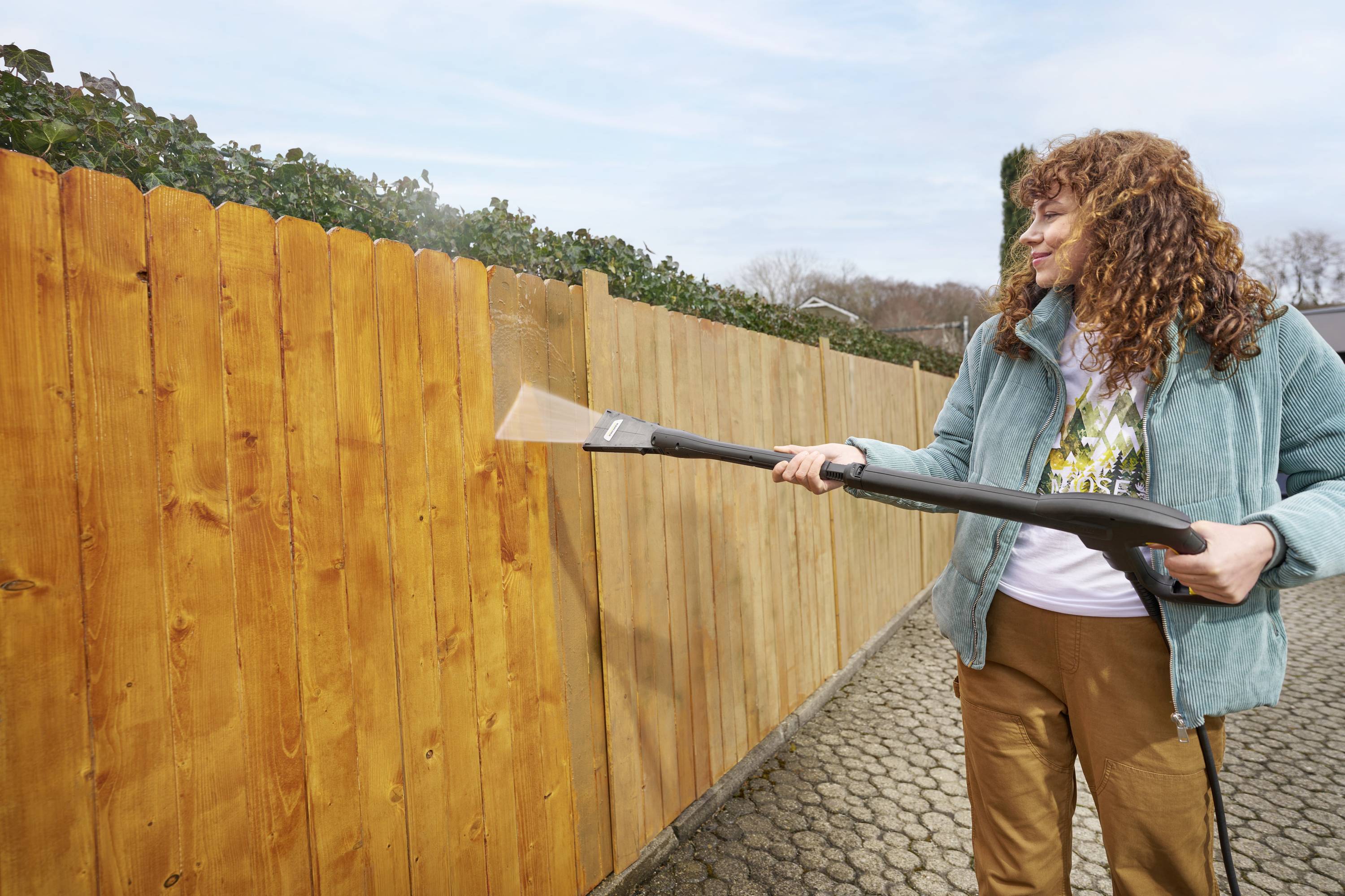 A person is cleaning a wooden fence with a pressure washer. The sky is clear, and the surroundings include a paved pathway.