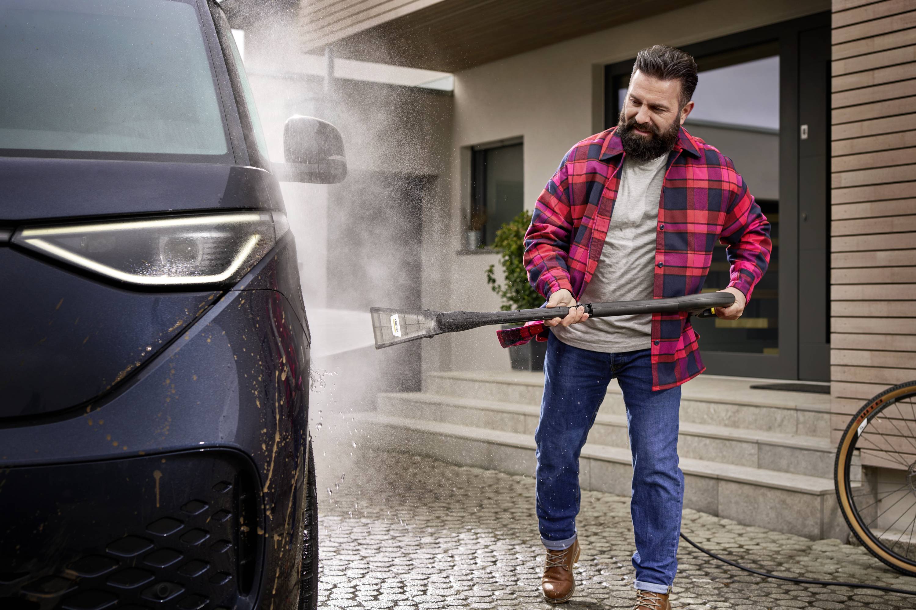 A man in a checked jacket is washing a car with a pressure washer on a paved driveway in front of a modern house.