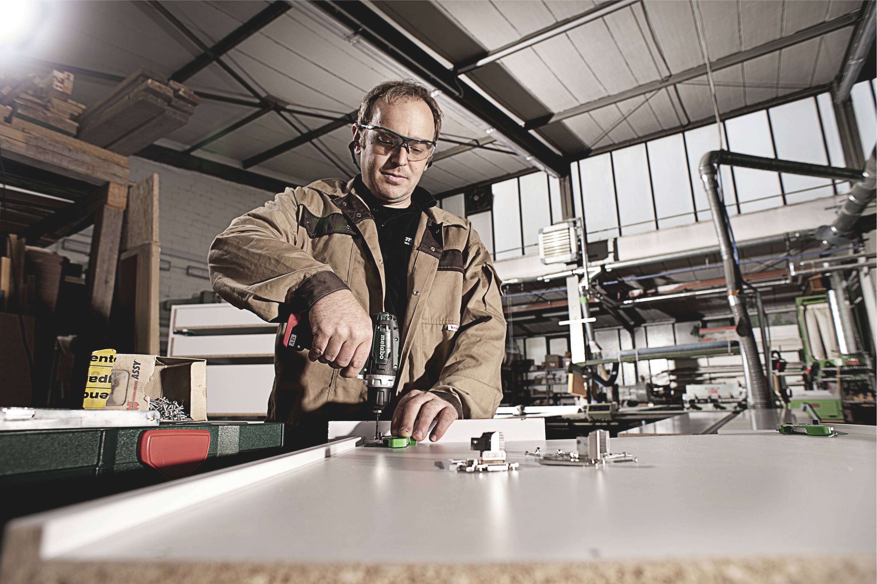A man is working in a workshop and using a drill, working on a workbench. Machines are visible in the background.