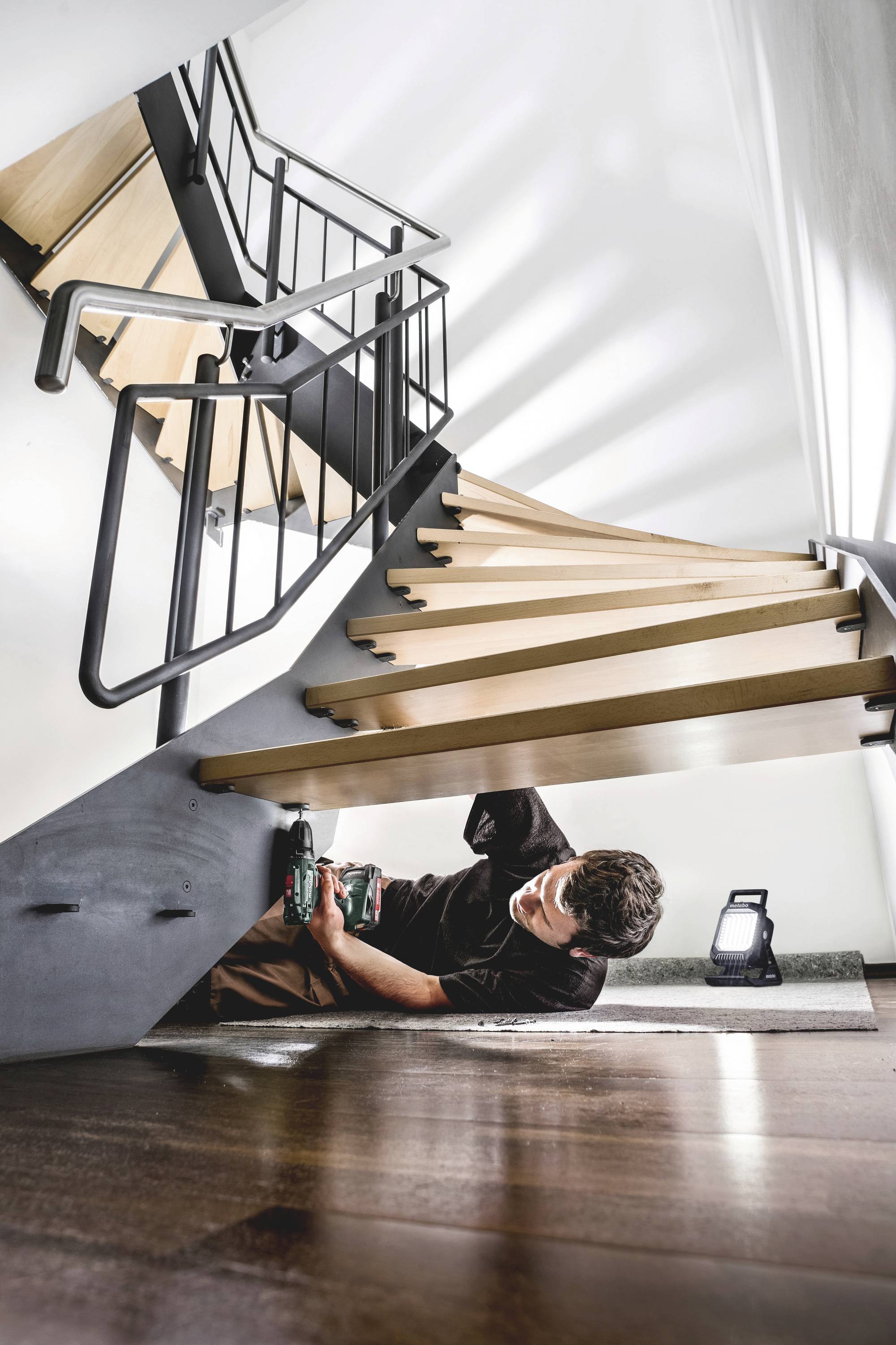 A person is repairing a wooden staircase from underneath using tools, lying on a wooden floor, with a small work lamp beside them.