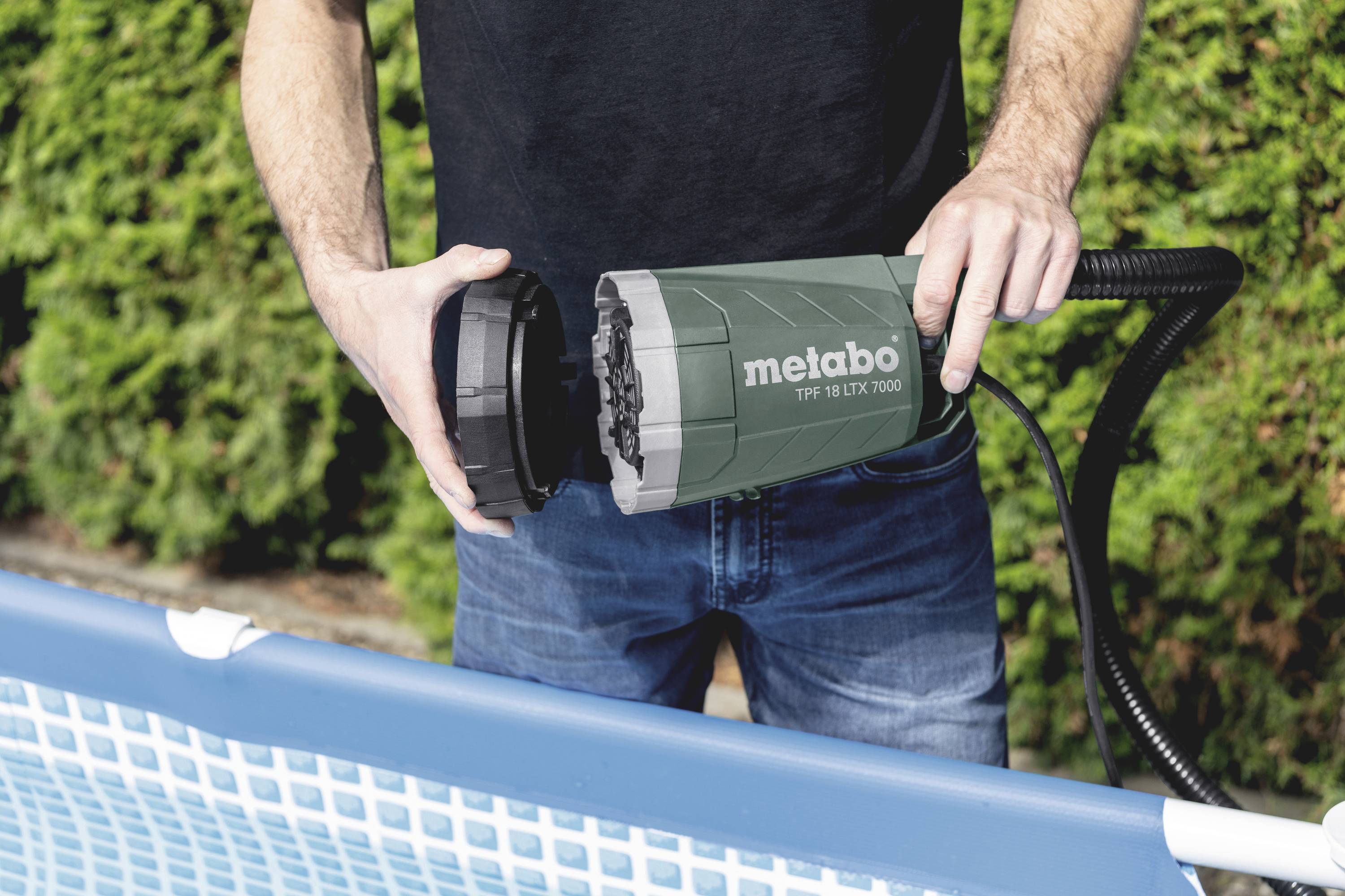 A person is holding a Metabo pool pump at the edge of a swimming pool. Background: green hedge.